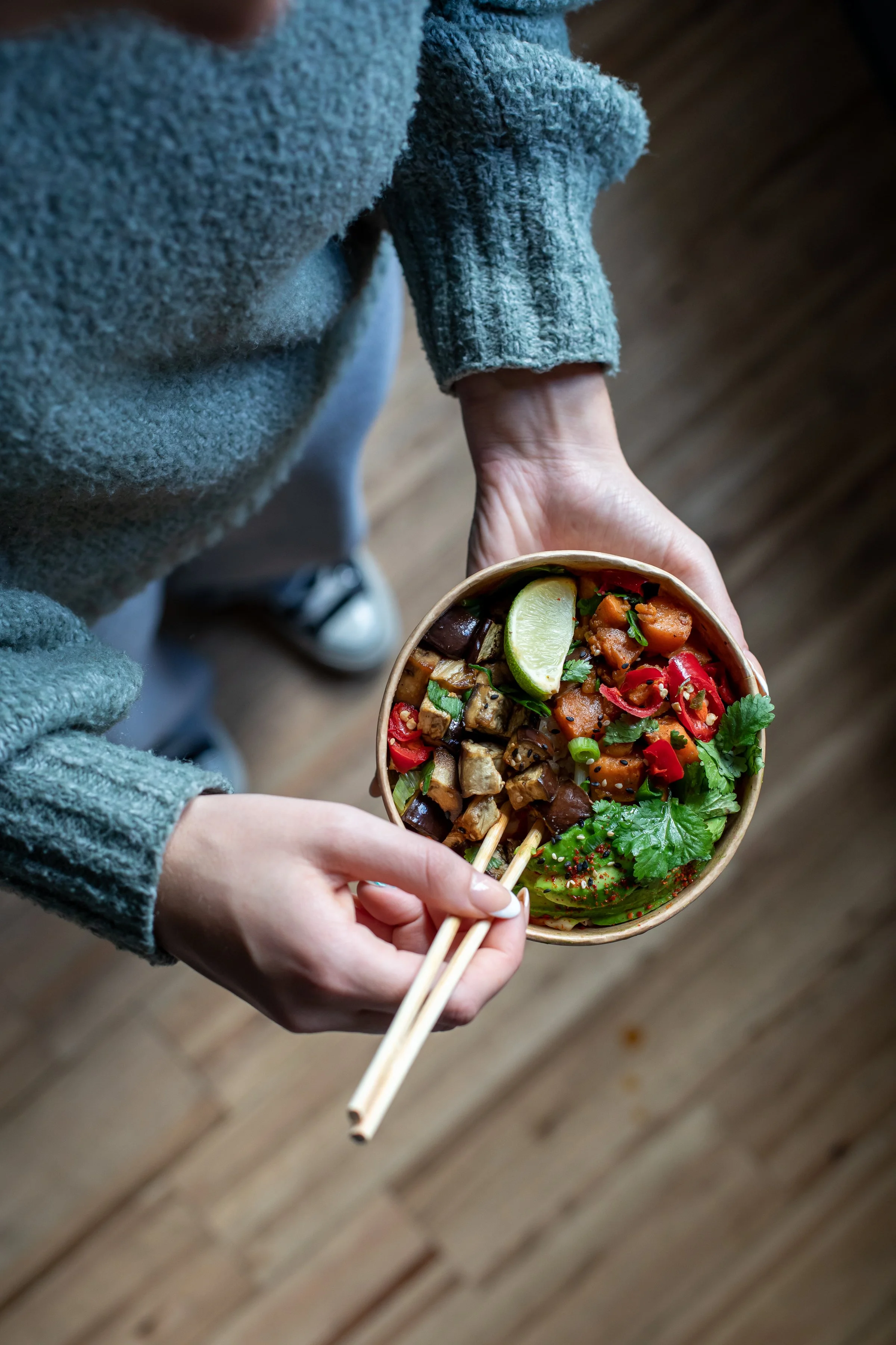 Person holding a bowl of assorted Asian-style vegetables and grilled skewers with chopsticks, a lime wedge on top, on a wooden floor.
