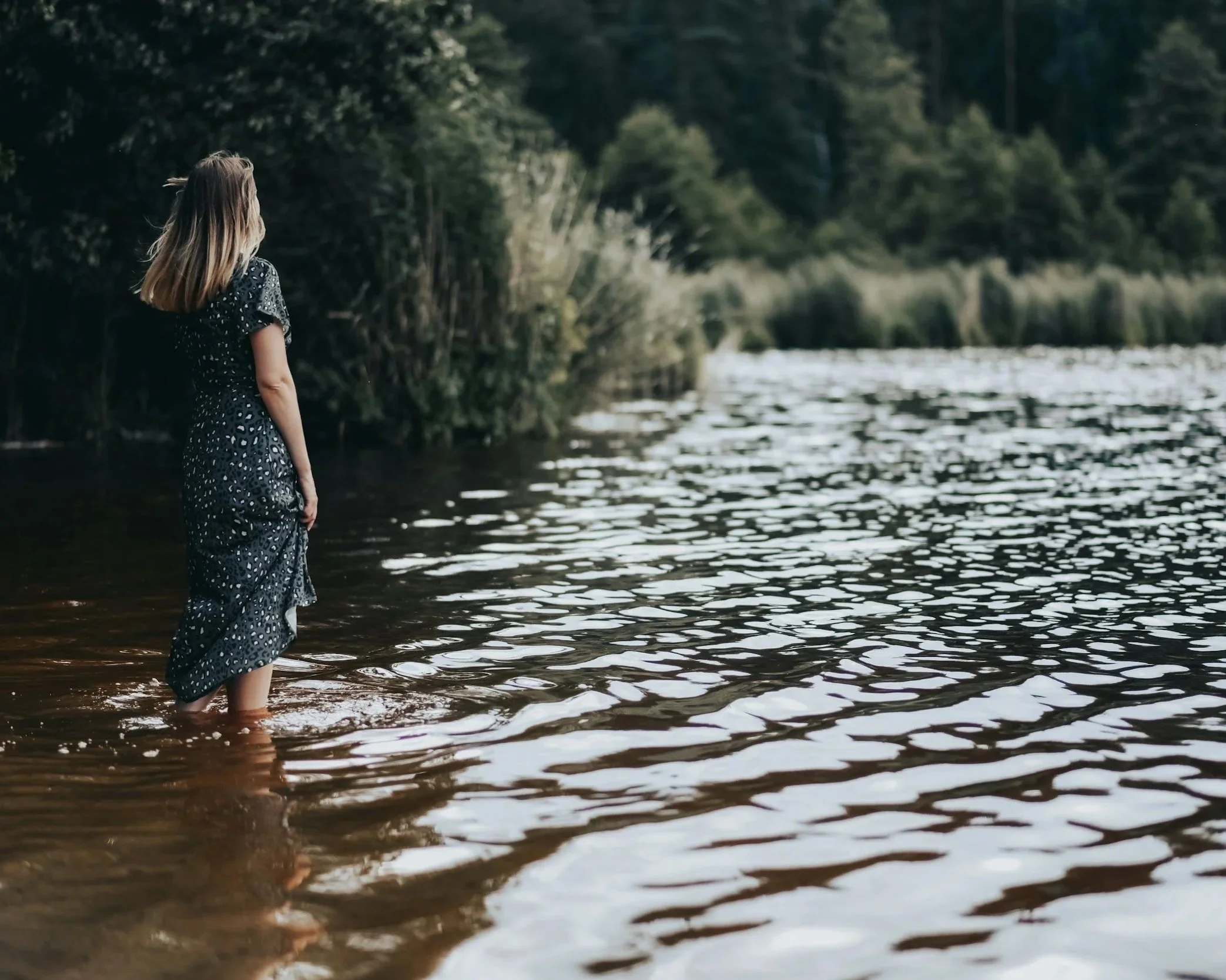 A woman with long hair in a dark dress standing in shallow water at a lake or river, surrounded by trees and nature.