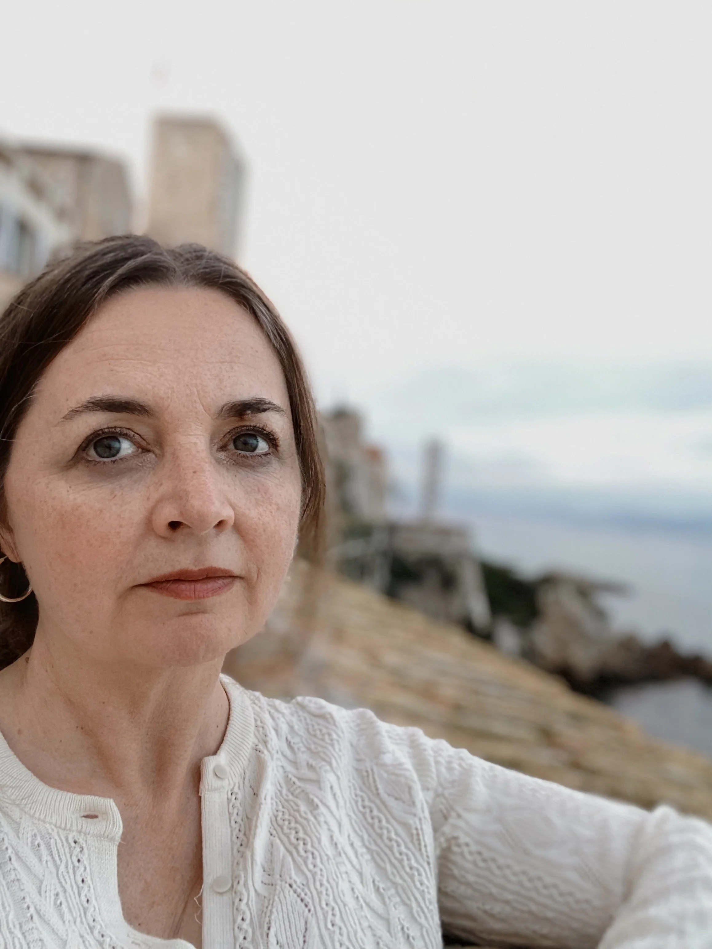A woman with short brown hair and blue eyes outdoors near the ocean, with buildings and rocks in the background.
