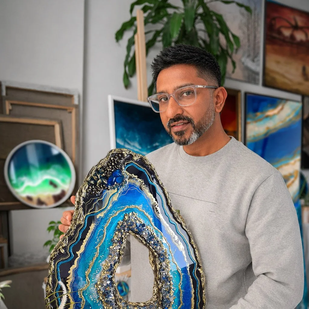 A man with glasses and a beard holding an abstract blue and gold artwork at an art gallery.
