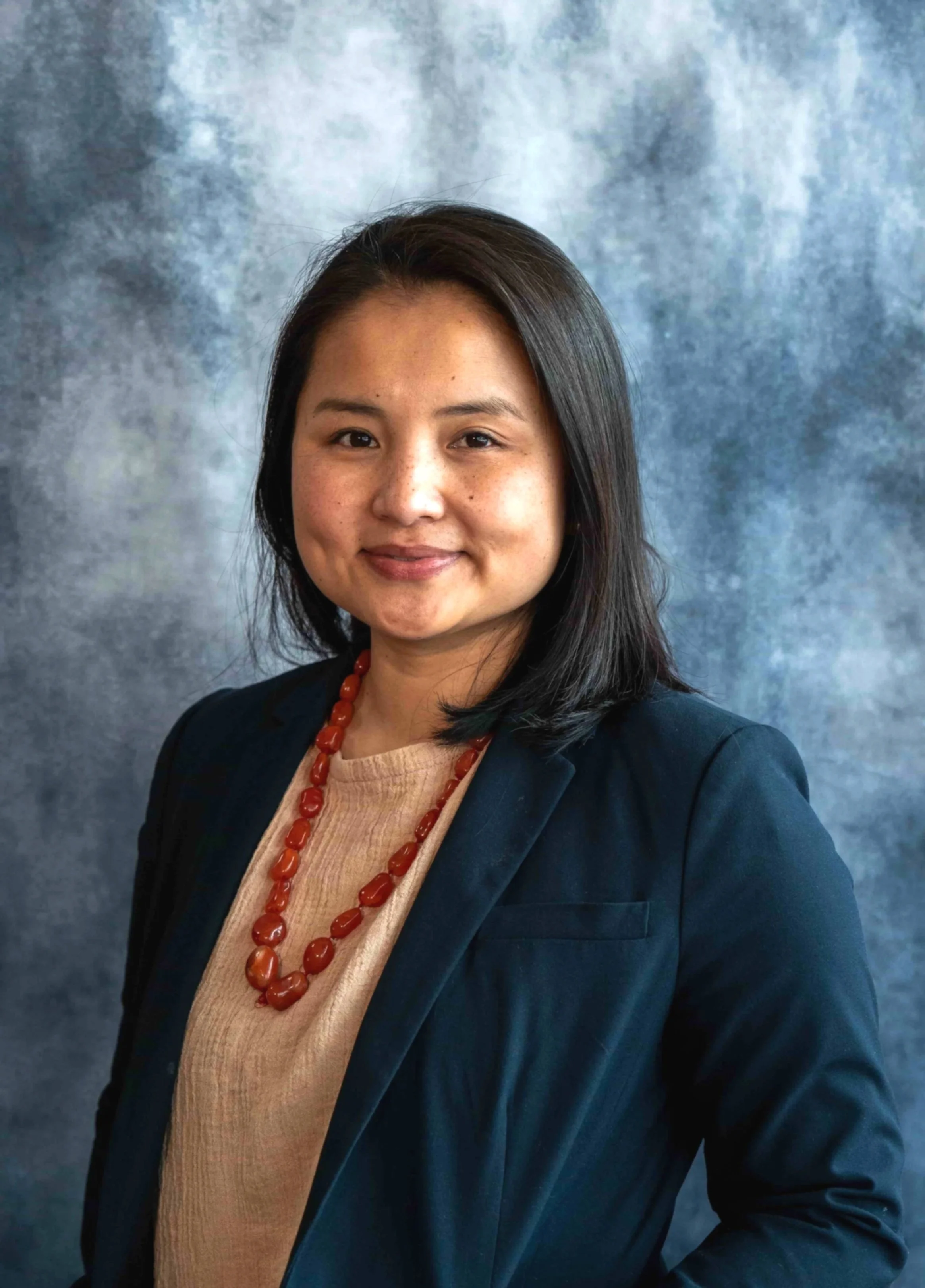 A woman with shoulder-length black hair, wearing a navy blazer, beige top, and a red beaded necklace, posing in front of a textured blue background.