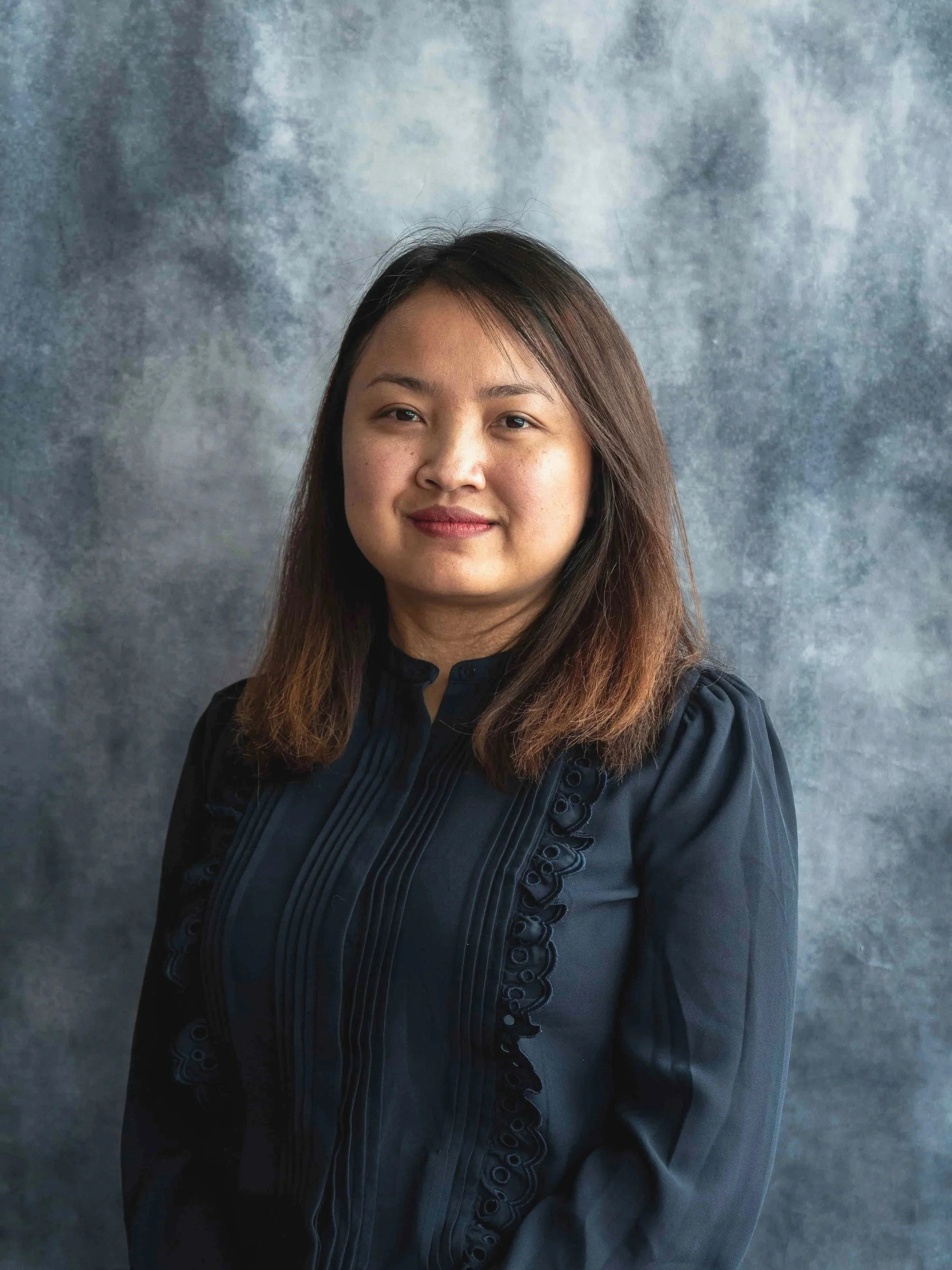 A woman with medium-length brown hair and a subtle smile, wearing a dark blouse with embroidered details, standing against a textured gray background.