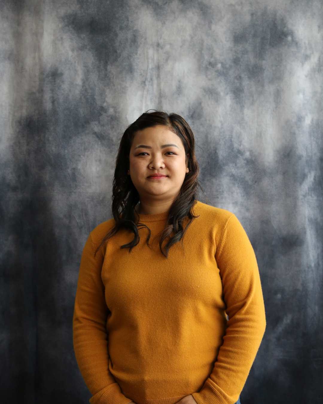 A woman with dark wavy hair wearing a mustard yellow sweater stands in front of a mottled gray background, looking at the camera with a slight smile.