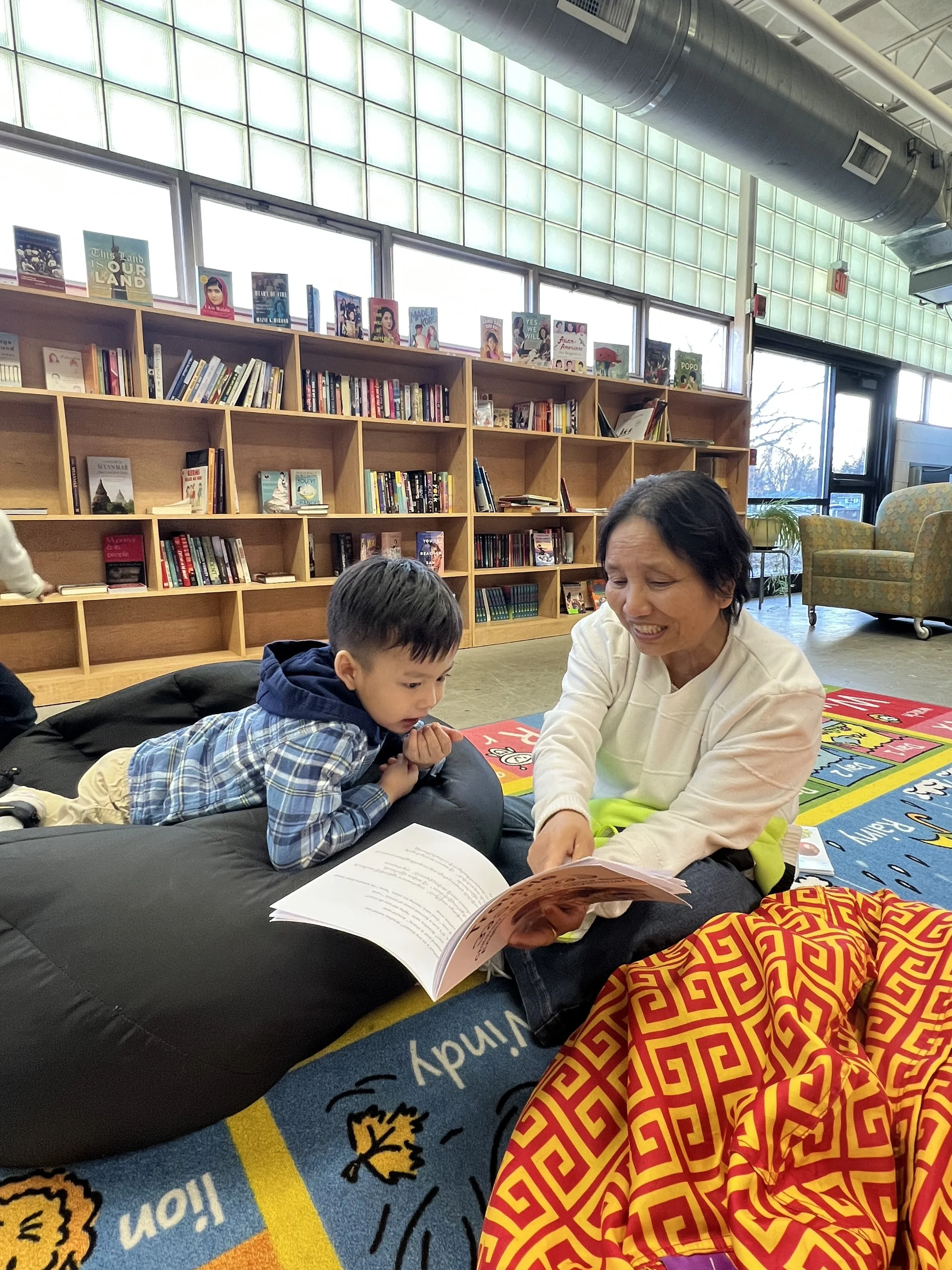 A woman and a young boy are sitting on the floor inside a bookstore or library, with the woman reading a book to the boy. They are sitting on a colorful children's rug, with bookshelves filled with books in the background.