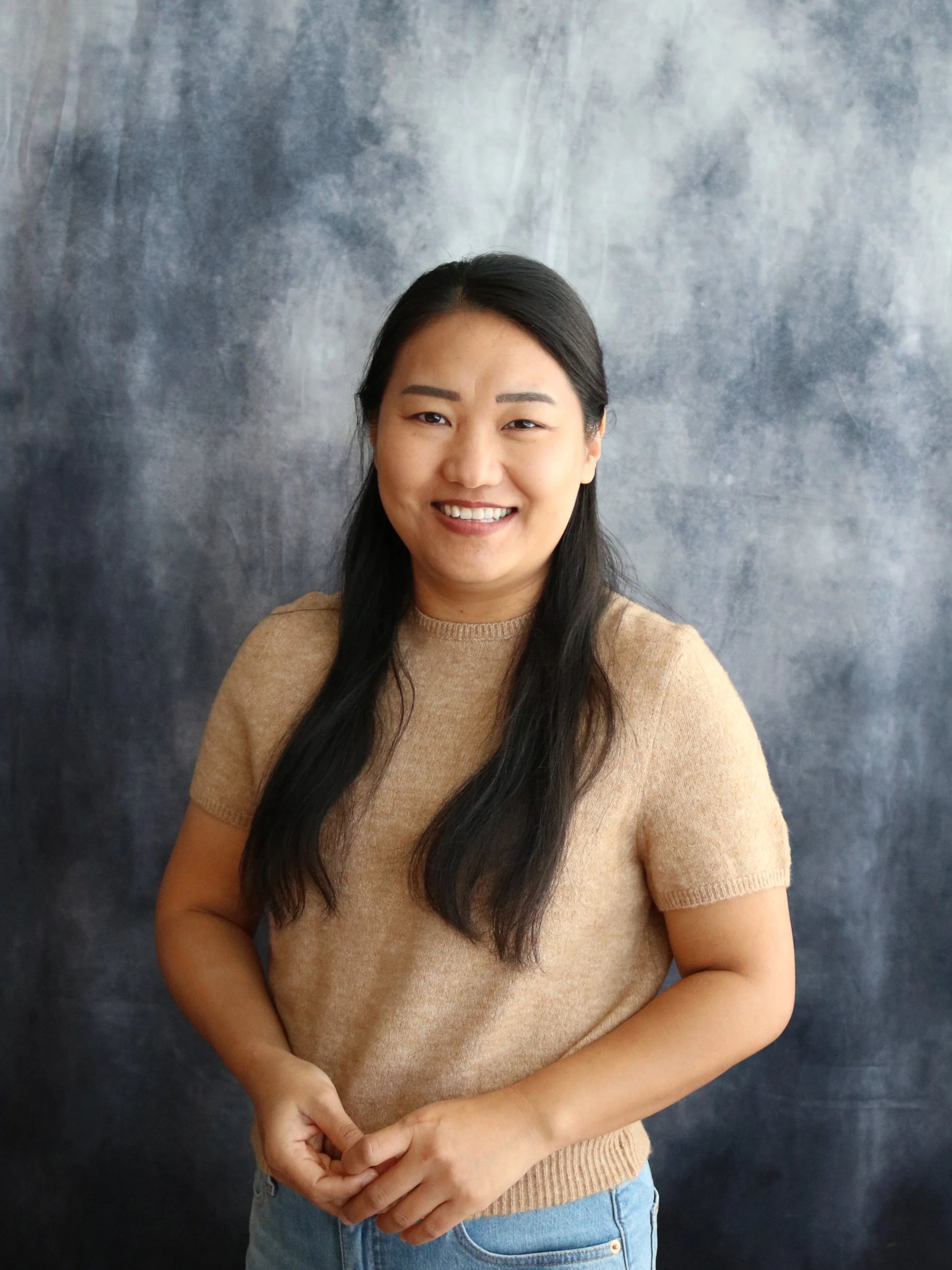 A woman with long dark hair smiling and standing against a gray textured background, wearing a tan short-sleeved sweater and blue jeans.