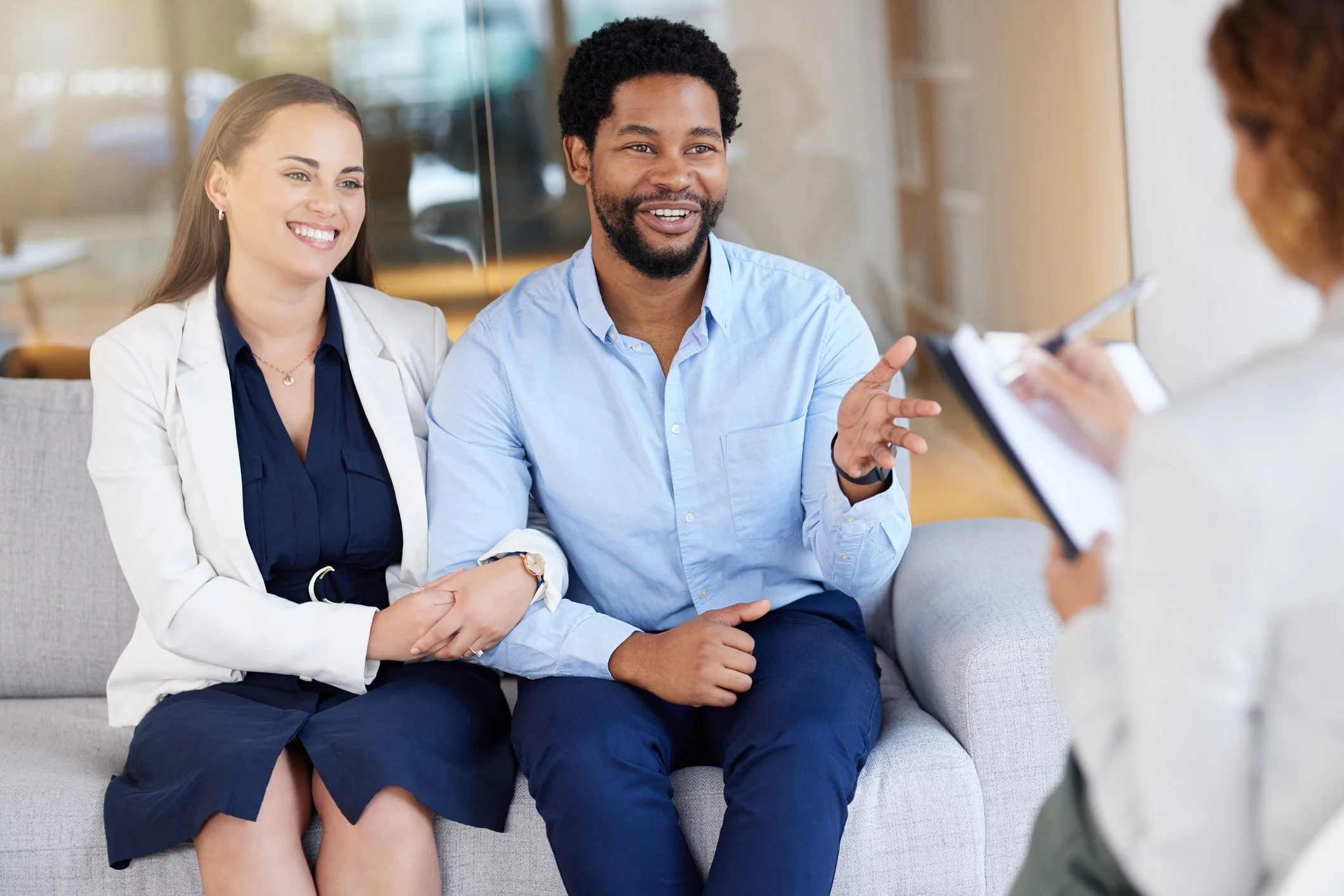 A couple holding hands and smiling while sitting together on a couch during a couples therapy session, representing engaged and motivated partners at AtReef Therapy in Cambridge, MA.