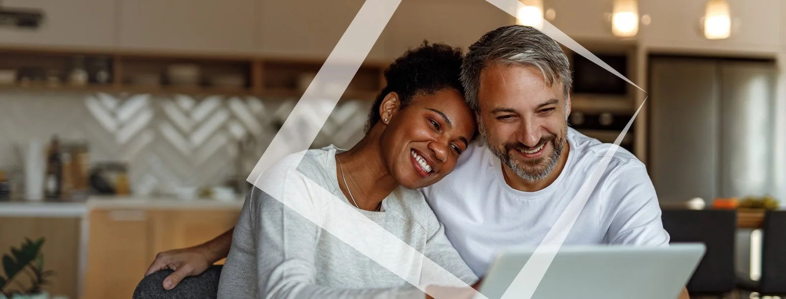 A smiling couple, sitting close together, looking at a laptop in a cozy kitchen.