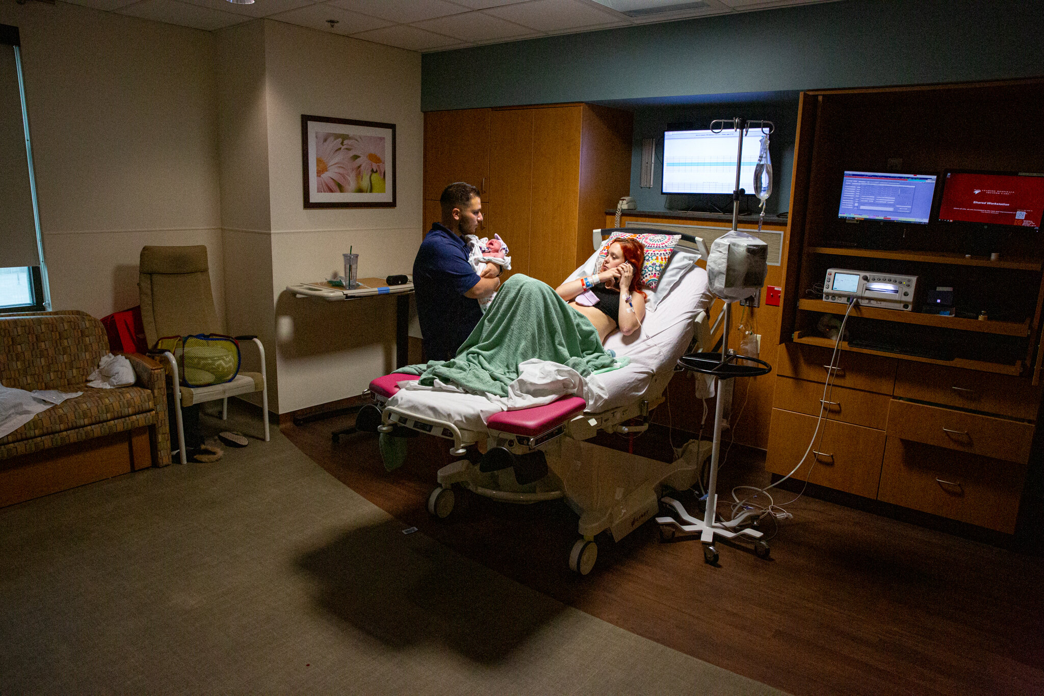 Hospital room with a woman lying in bed and man holding baby standing next to her