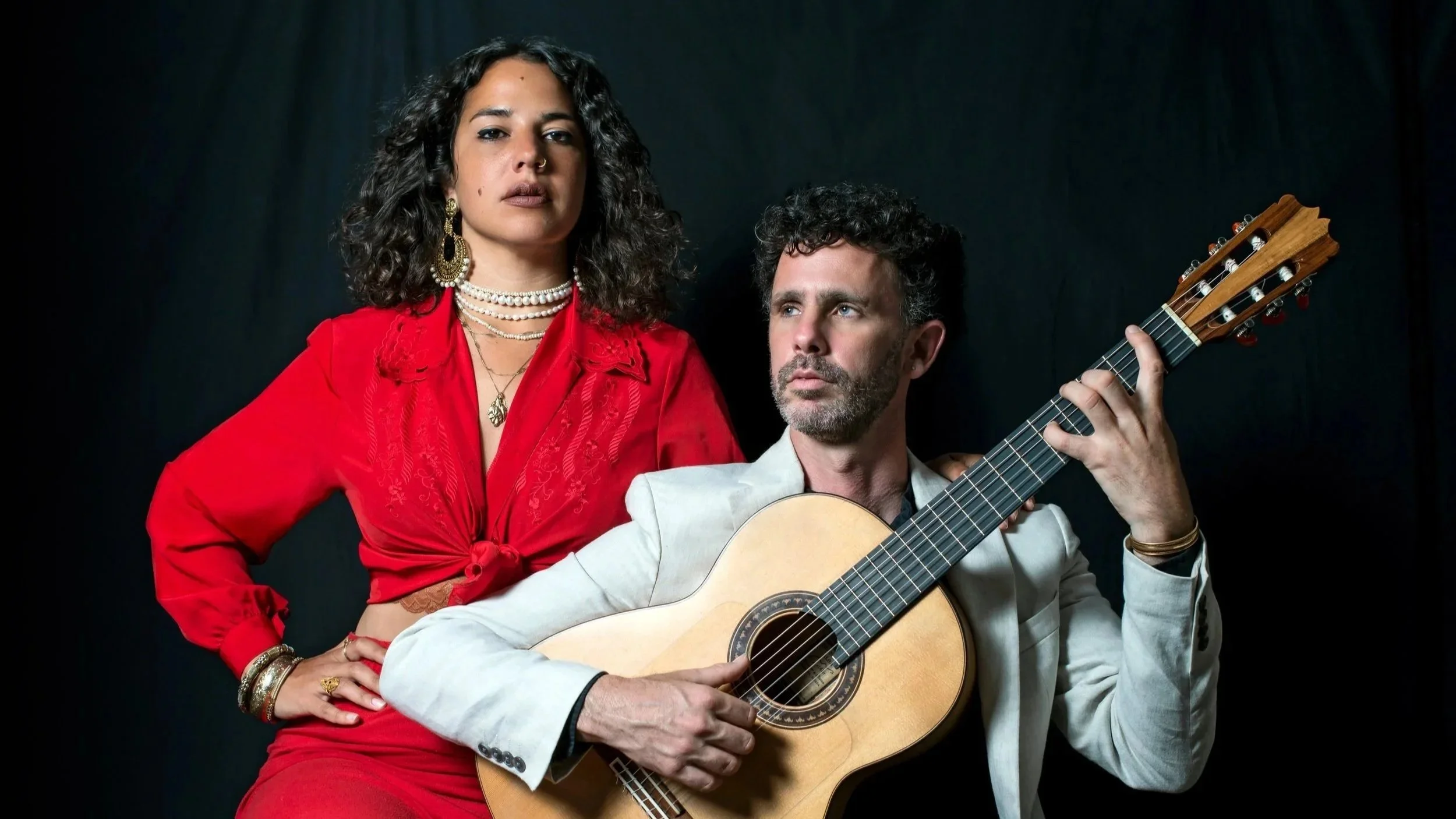LALA Tamar in a red outfit, looking at the camera and standing behind Ofer Ronen in a white jacket, holding a guitar.