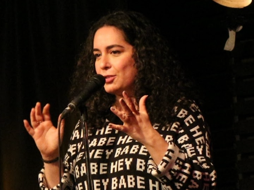 Ronit Rubinstein wearing a shirt that says Hey Babe, standing and speaking at a microphone. Photo by Matthew McLaren.