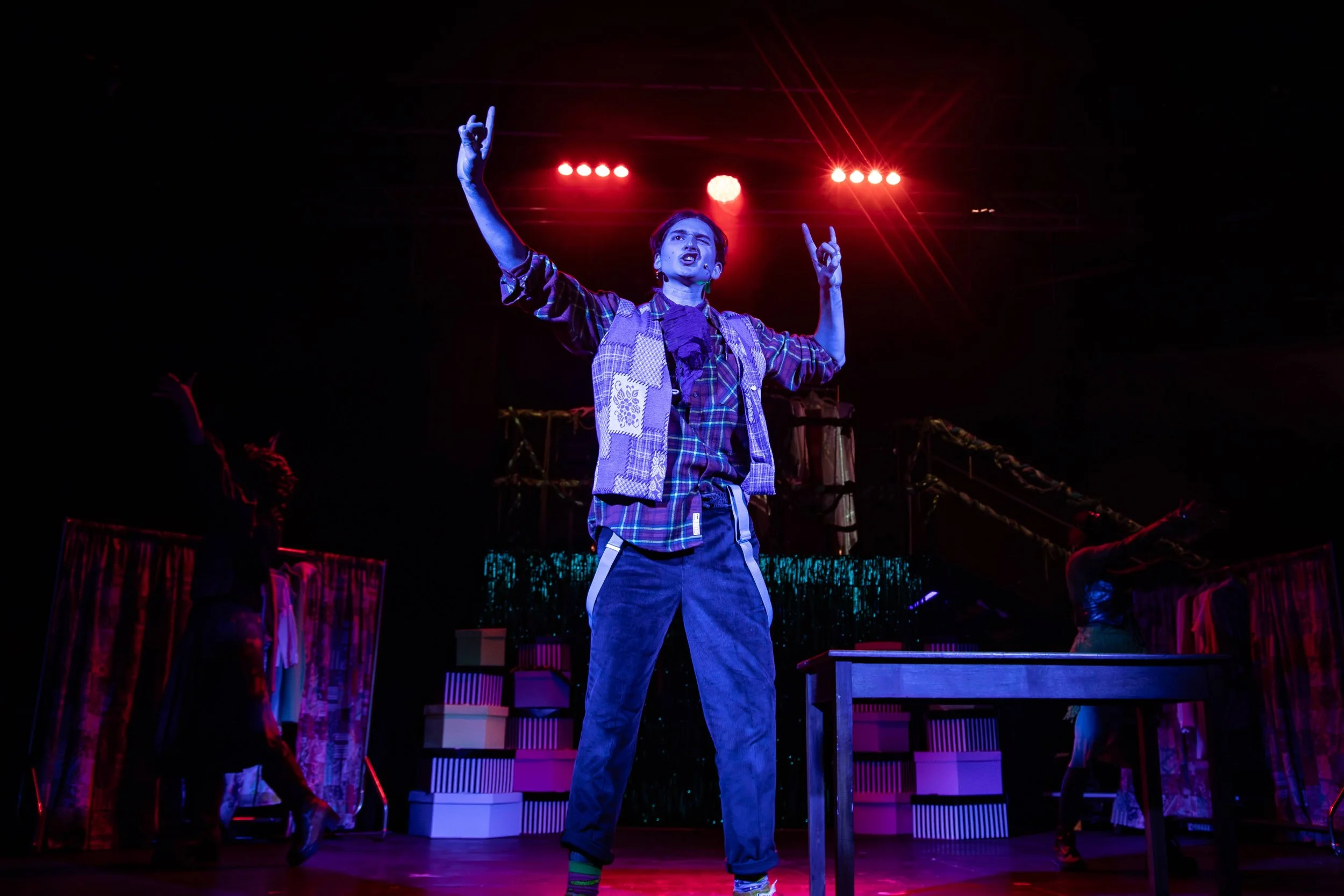 Yankl, bathed in blue light, standing center stage, wearing a patchwork vest, plaid, and suspenders.