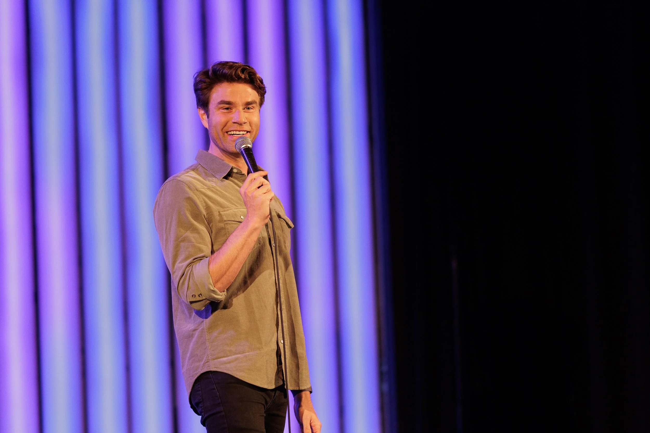 Matthew smiling broadly, wearing a khaki coloured button-up shirt while holding a microphone, in front of a background of purple lights.