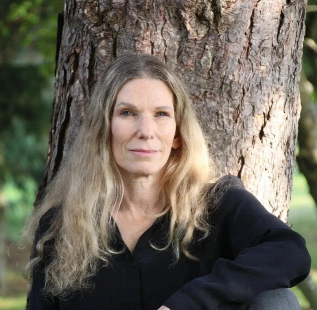Lilach with long blonde hair, in a black shirt, sitting with her back against a tree.