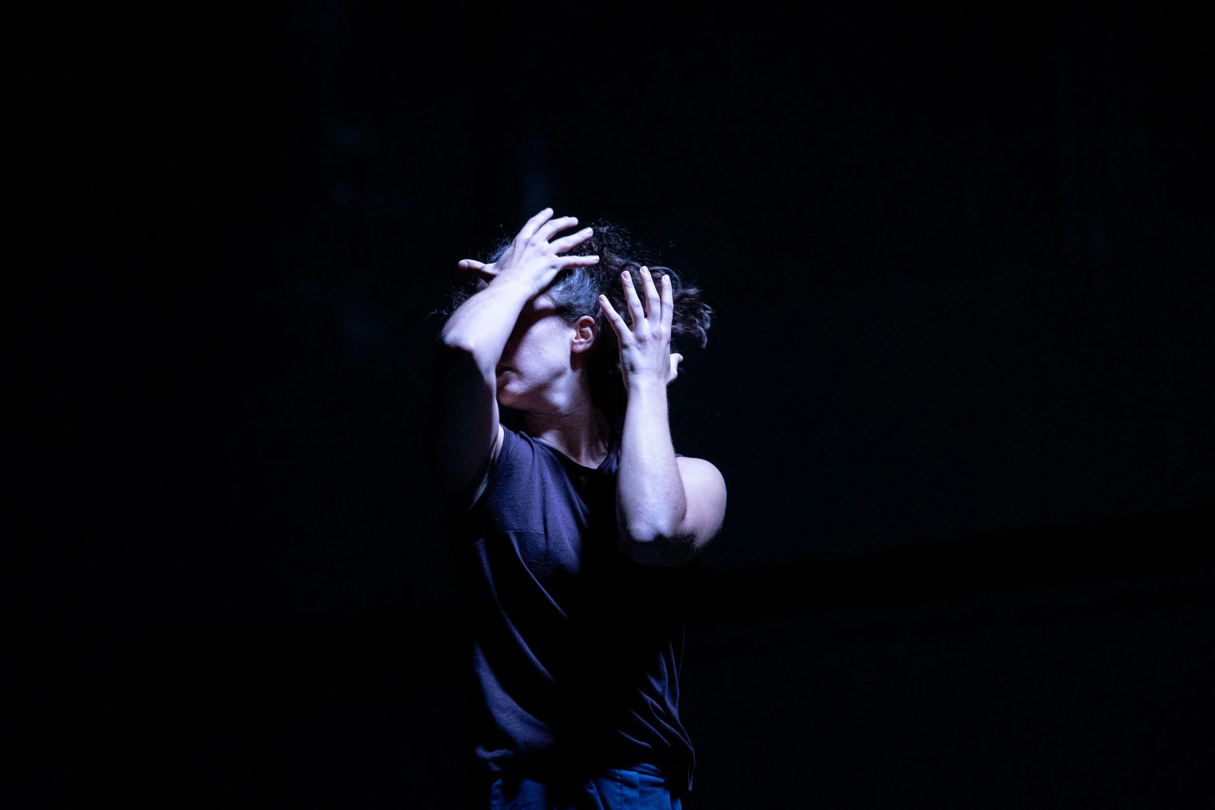 Amit, wearing a blue shirt, with her arms up in front of her face, in front of a dark background. Photo by Dor Pazuelo.