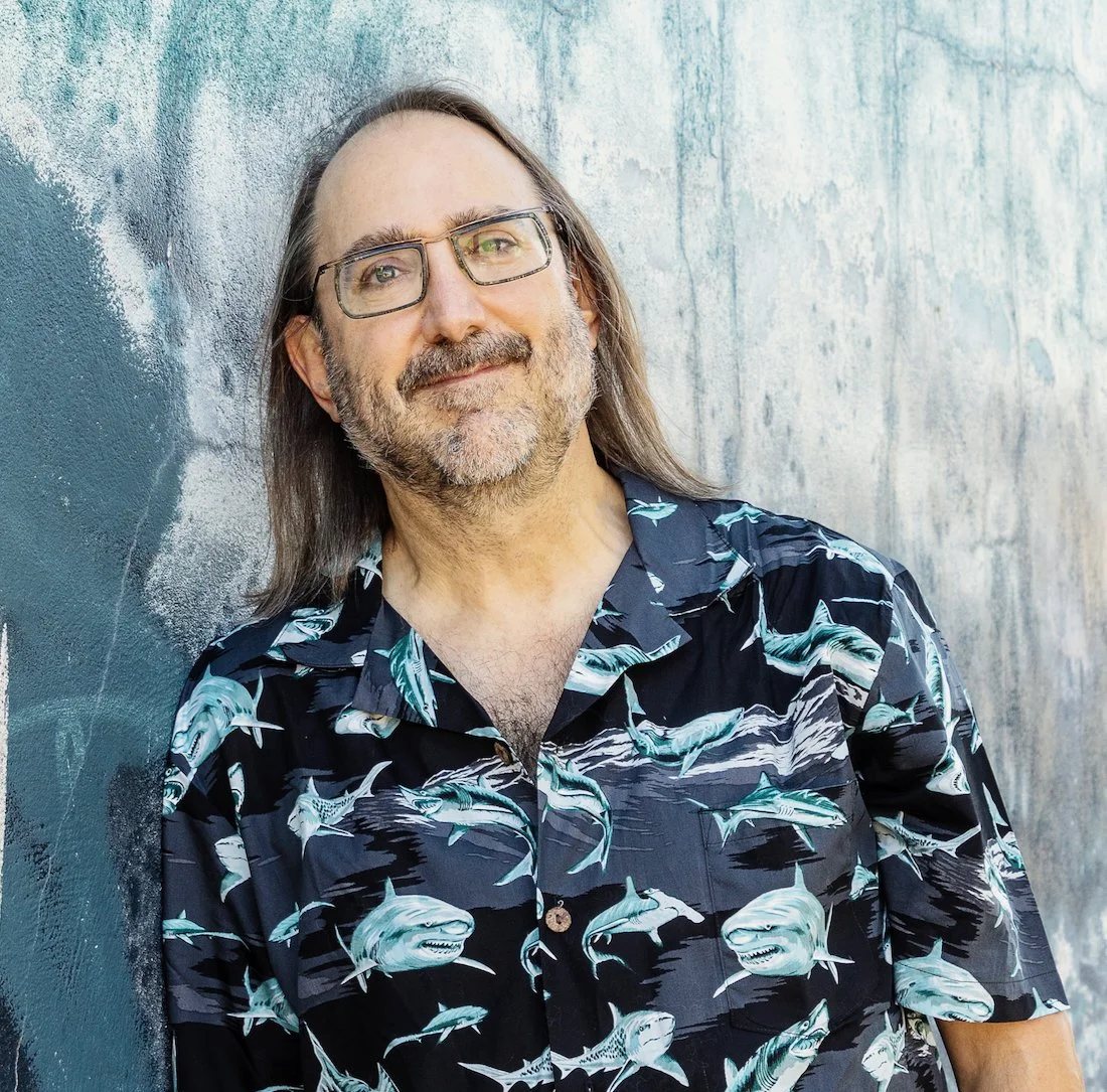 Mark with shoulder length hair, a short beard, square glasses, and in a button up covered in sharks.