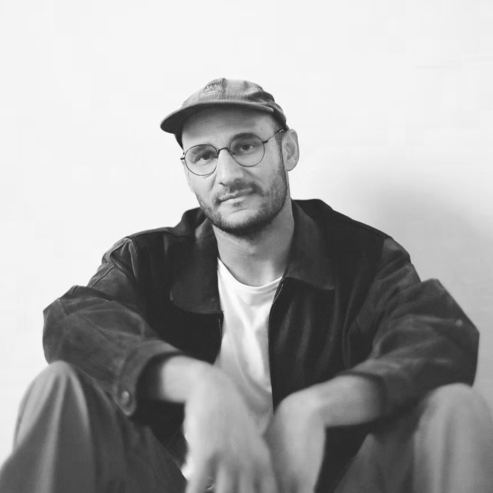 Black and white photo of a man with glasses, a beard, wearing a cap and a jacket, leaning forward with arms extended toward the camera, against a plain background.