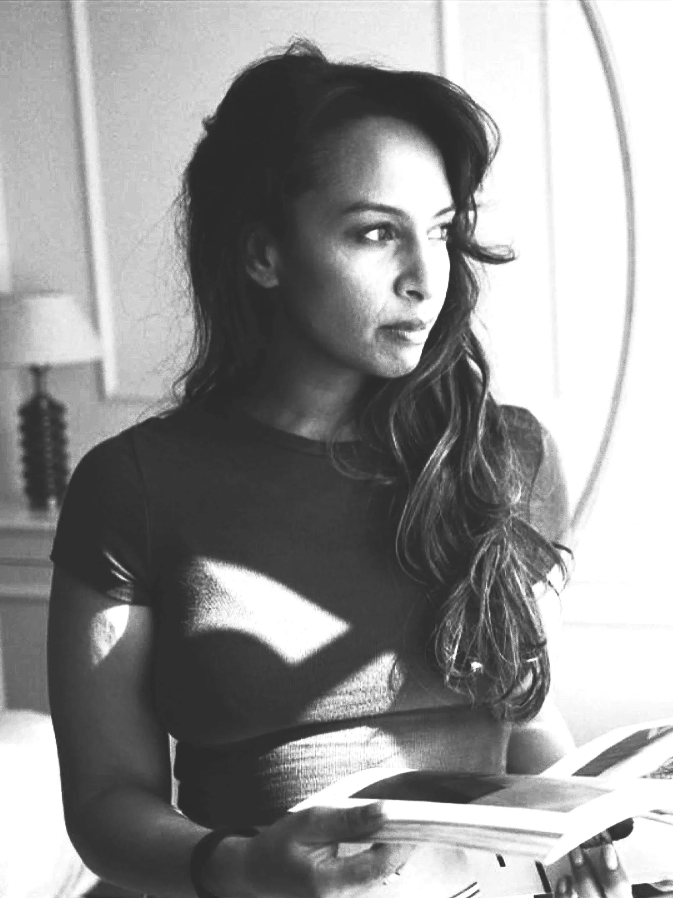 A woman with long wavy hair reading a book, sitting indoors near a lamp and a mirror.