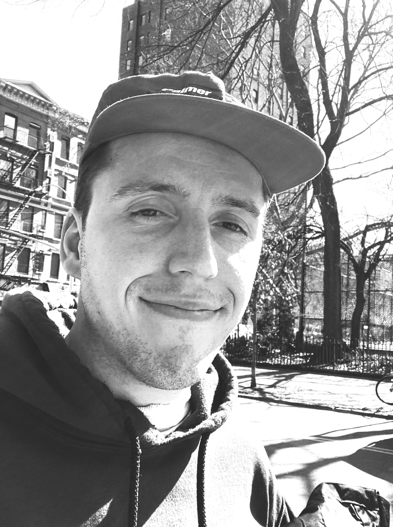 A young man smiling outdoors in a park on a sunny day, wearing a baseball cap and hoodie, with trees and buildings in the background.