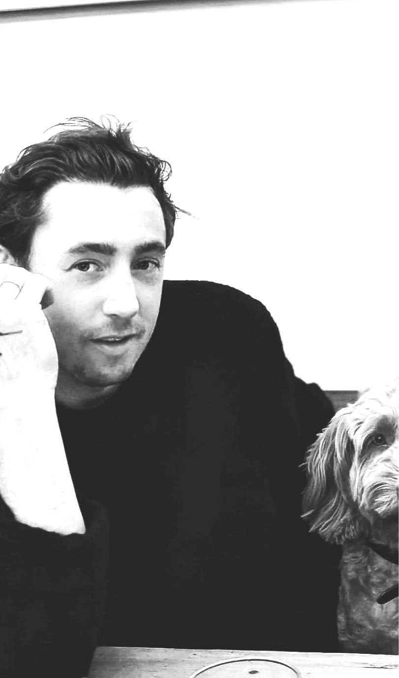 A black and white photo of a man with dark hair and eyebrows, wearing a dark sweater, sitting at a table with a small dog with curly fur, probably a cockapoo, sitting on his right side.