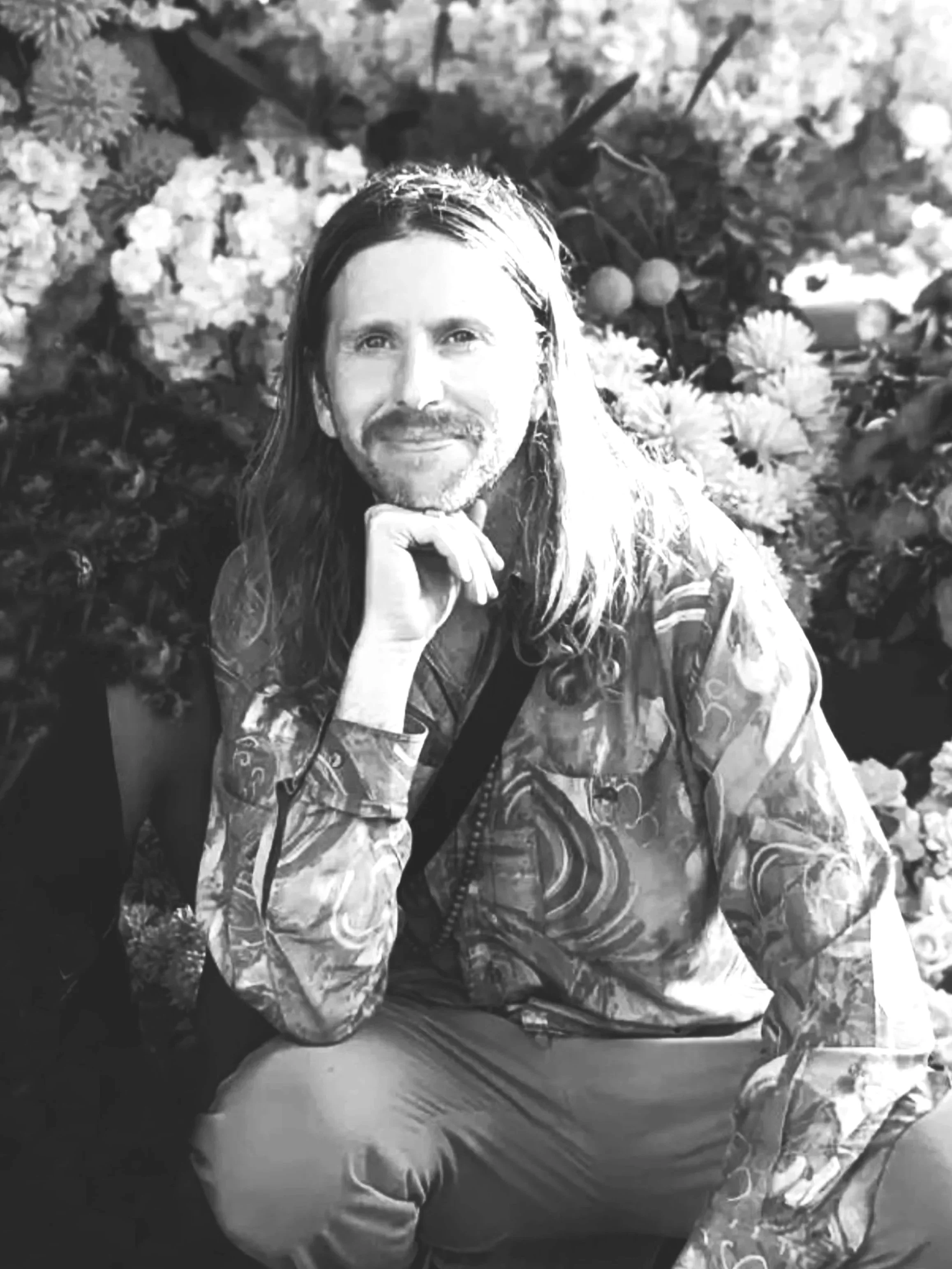 Black and white photo of a man with long hair and a beard, sitting in front of a background of flowers, wearing a patterned jacket and light-colored pants.