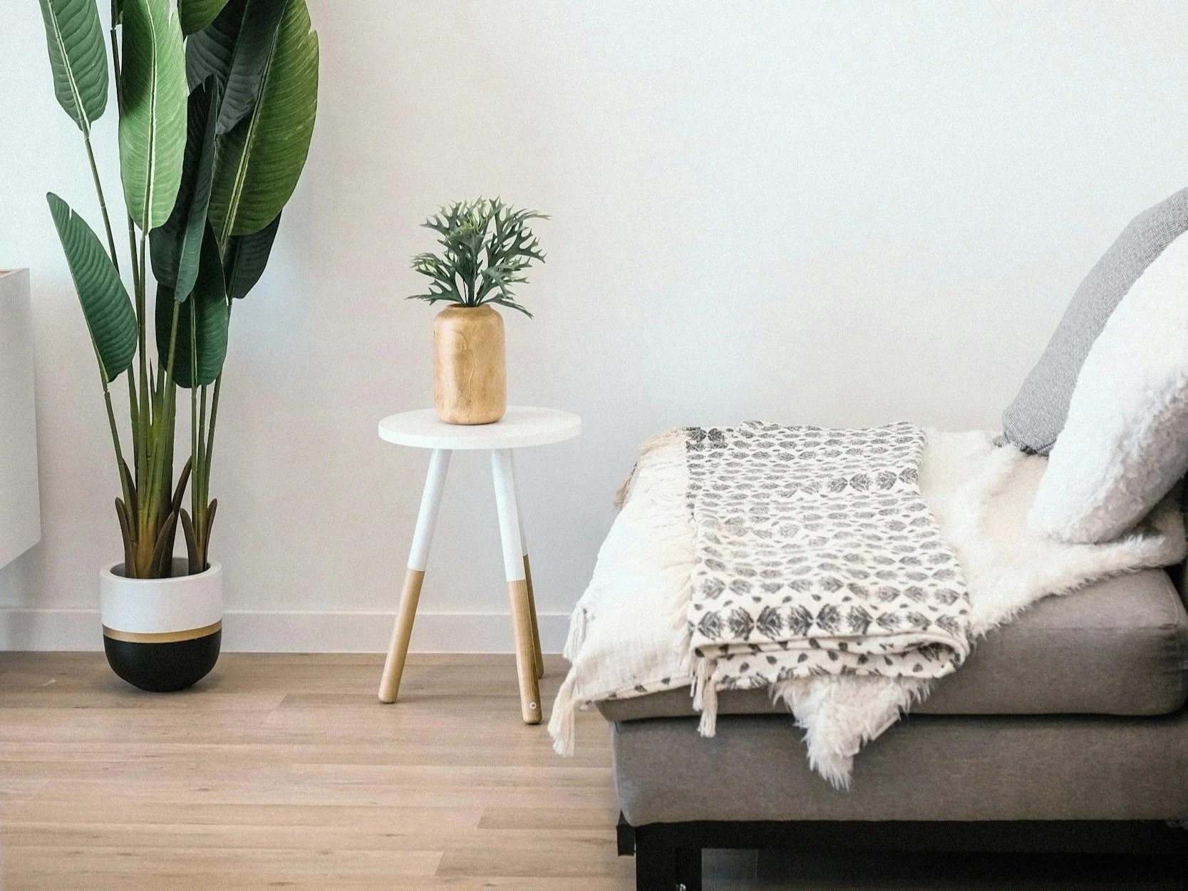 Minimalist bedroom corner with a large potted plant on the left, a small white side table with a vase and plant in the center, and a bed with a blanket and pillows on the right.