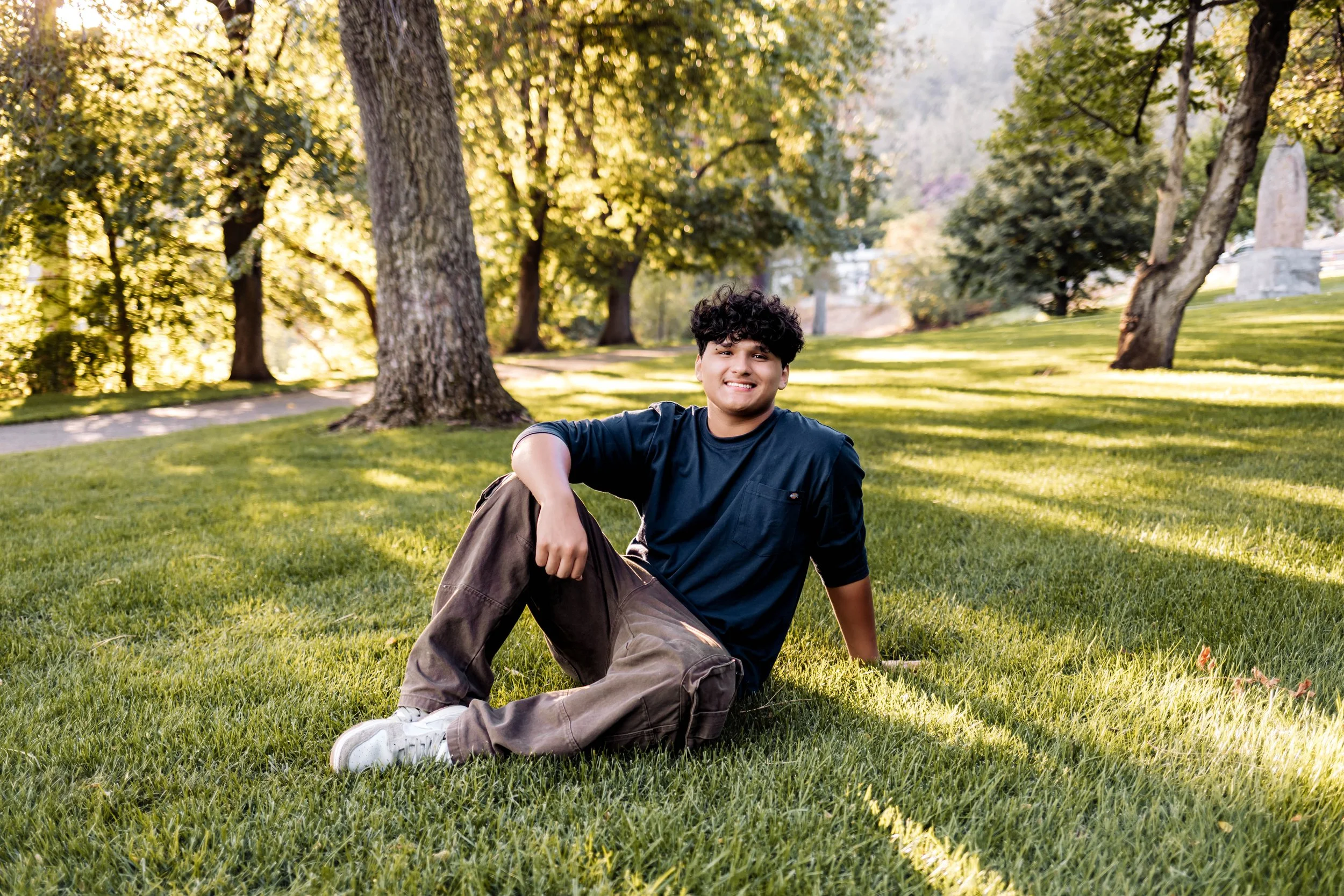 A young man sitting on a grassy park lawn, smiling, with trees and a pathway in the background during daytime.