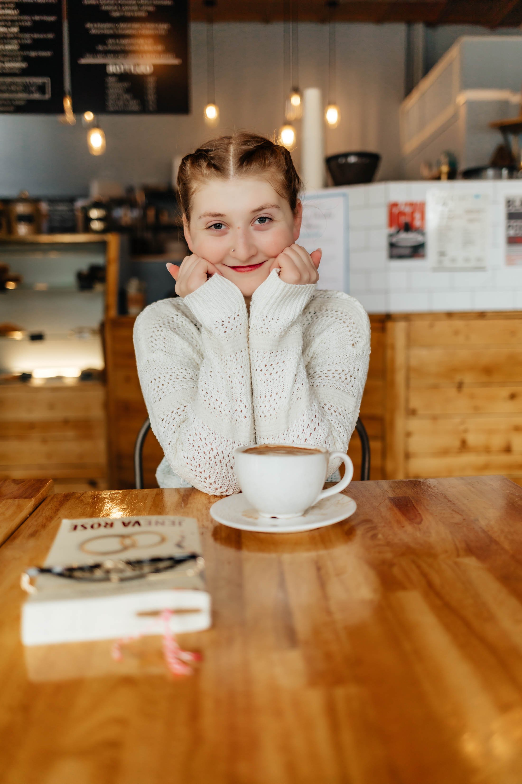 A young girl with braided hair and a nose piercing sitting at a wooden table in a cafe, smiling with her hands near her face, with a cup of coffee or hot chocolate in front of her and a small book or notebook on the table.