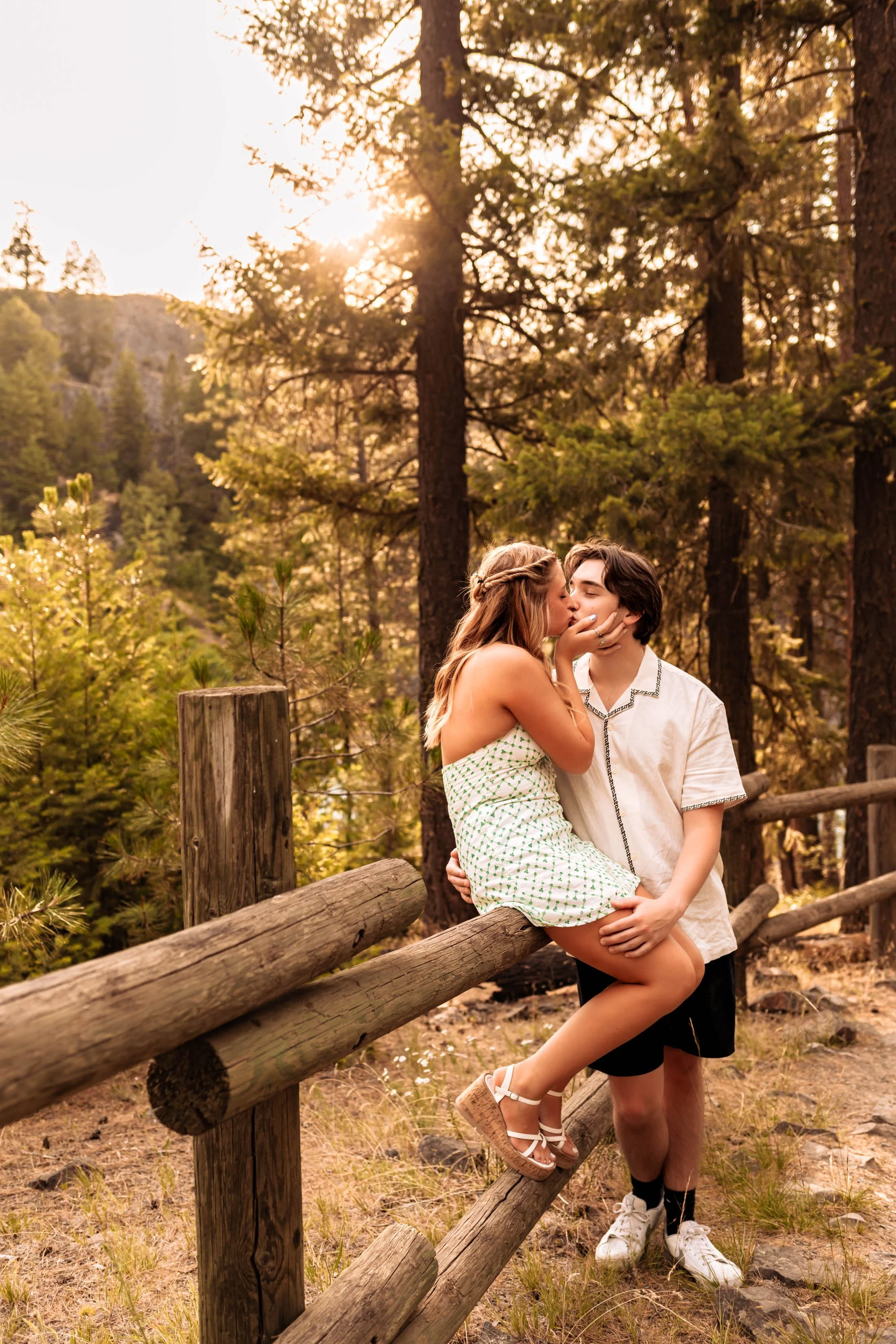 A young man in a white T-shirt and jeans standing outdoors near a tree, with a river and trees in the background.