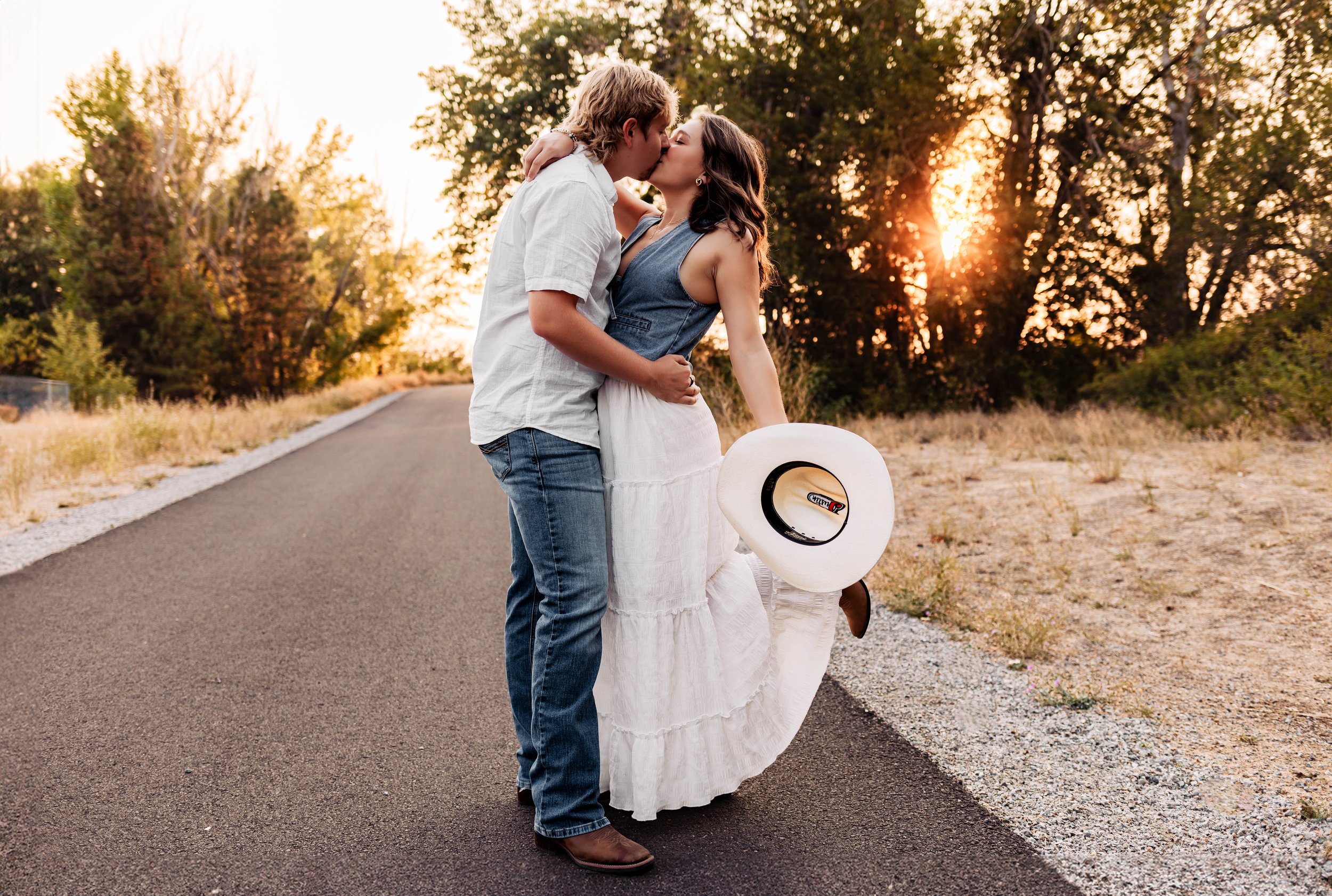 Pregnant woman in pink lace dress standing in a grassy field during sunset, gently holding her belly and looking down.