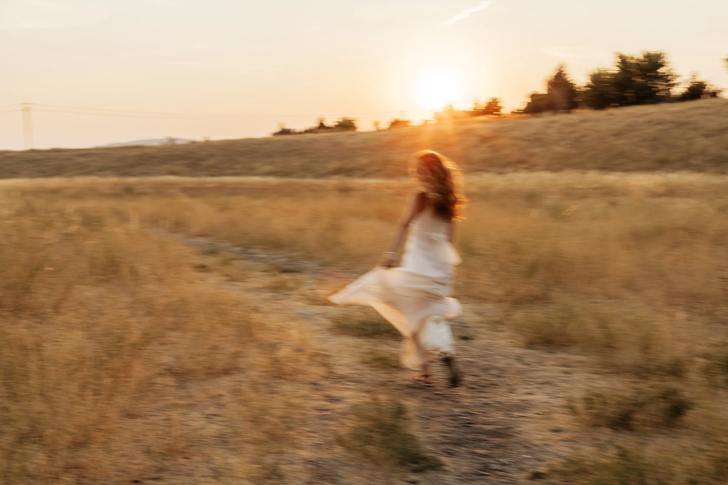A young woman with long, curly hair sitting on the ground in a field of small flowers at sunset, smiling and looking off to the side.