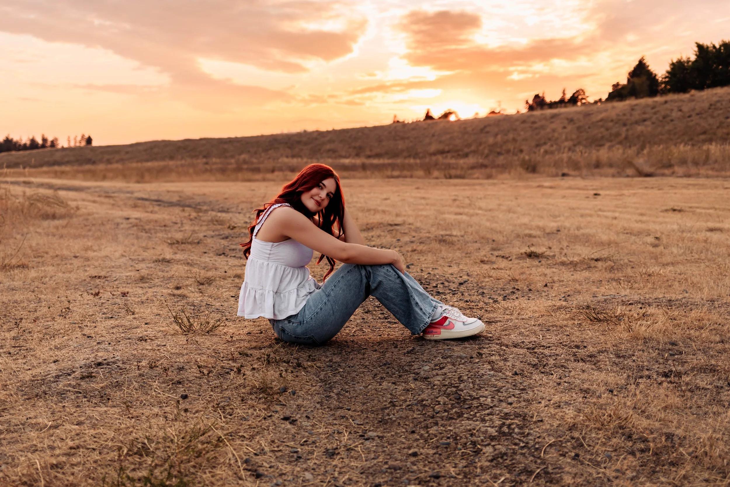 A young woman with long, curly hair sitting on the ground in a field of small flowers at sunset, smiling and looking off to the side.