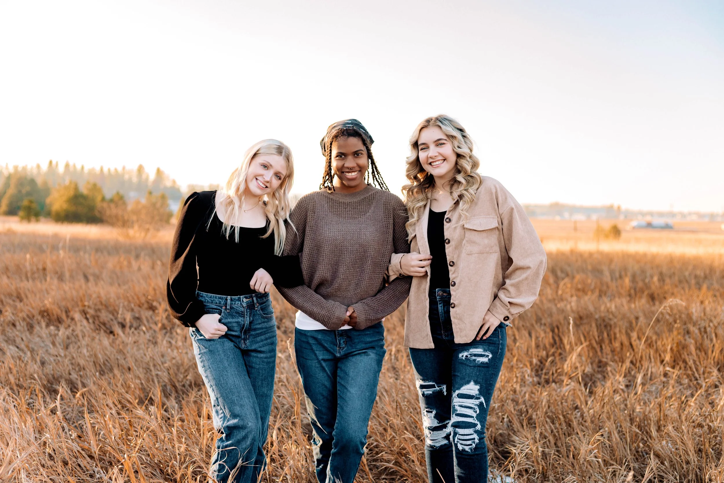 Three young women standing arm-in-arm in a field of tall grass at sunset, smiling at the camera.