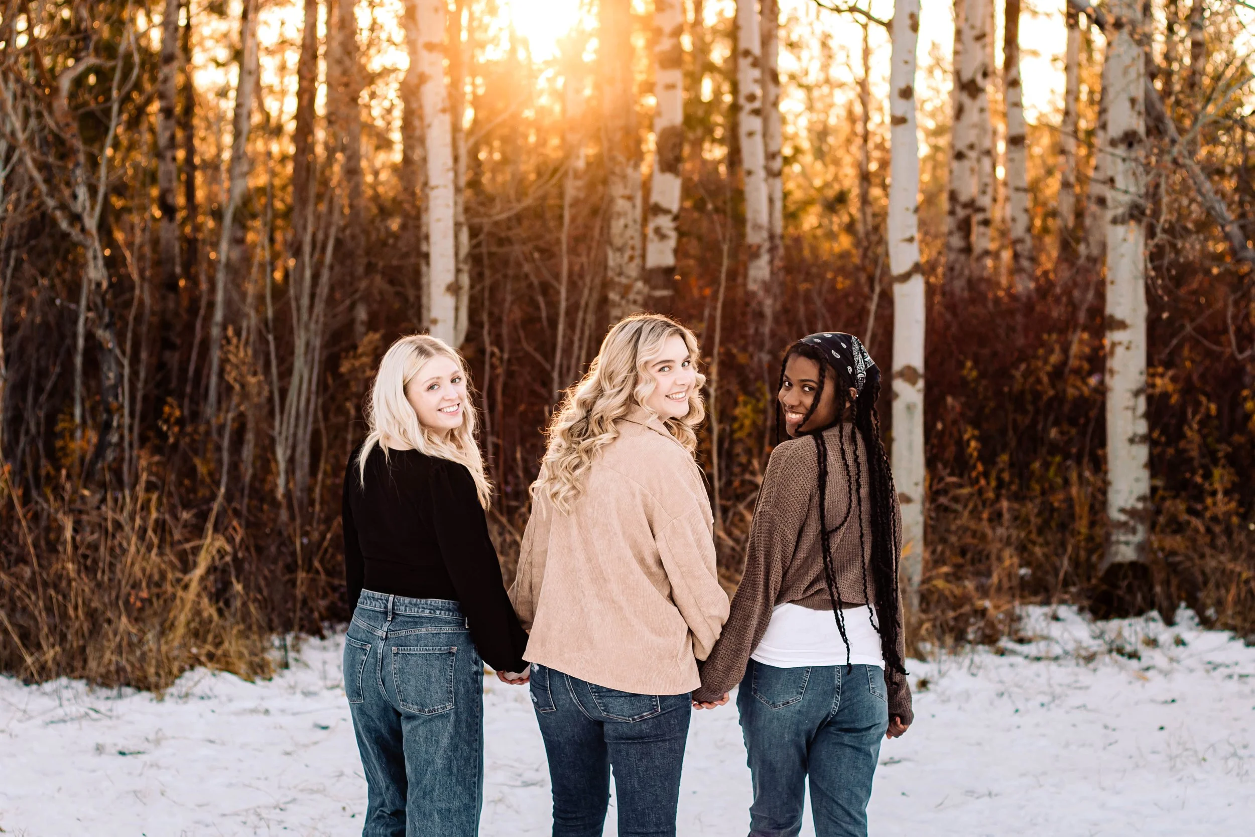Three young women holding hands and smiling in a snowy forest at sunset.