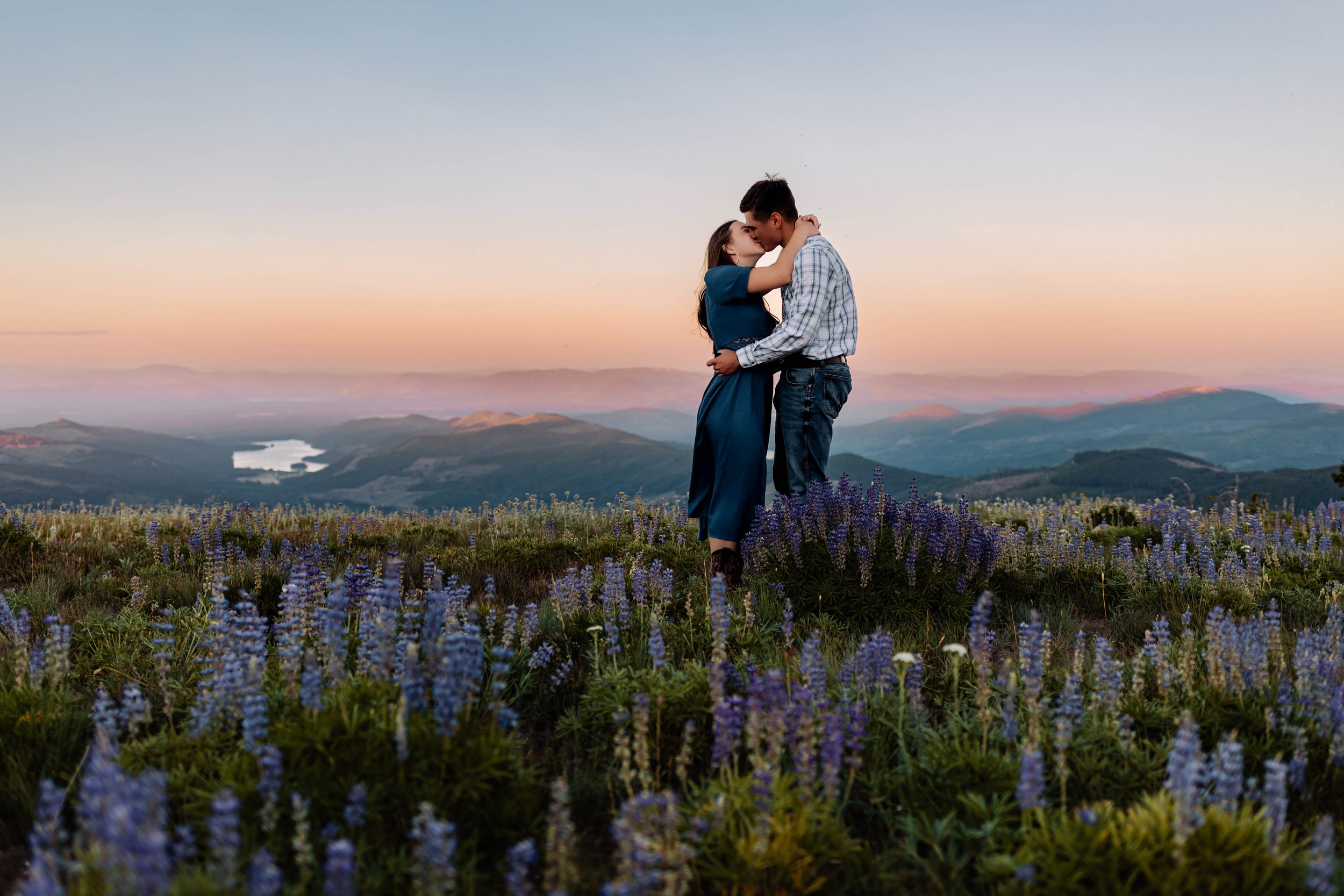 A couple kissing on a hilltop filled with purple and white wildflowers, with a scenic mountain landscape and lake in the background during sunset.