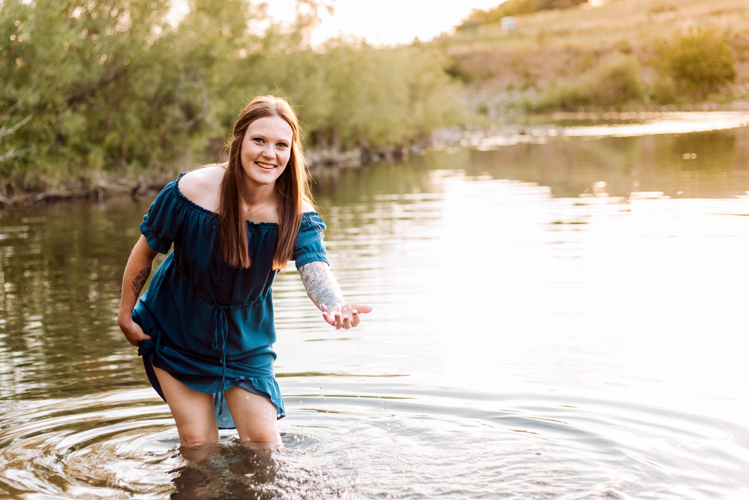 A woman in a blue dress wading in a river at sunset, smiling and reaching out her hand.