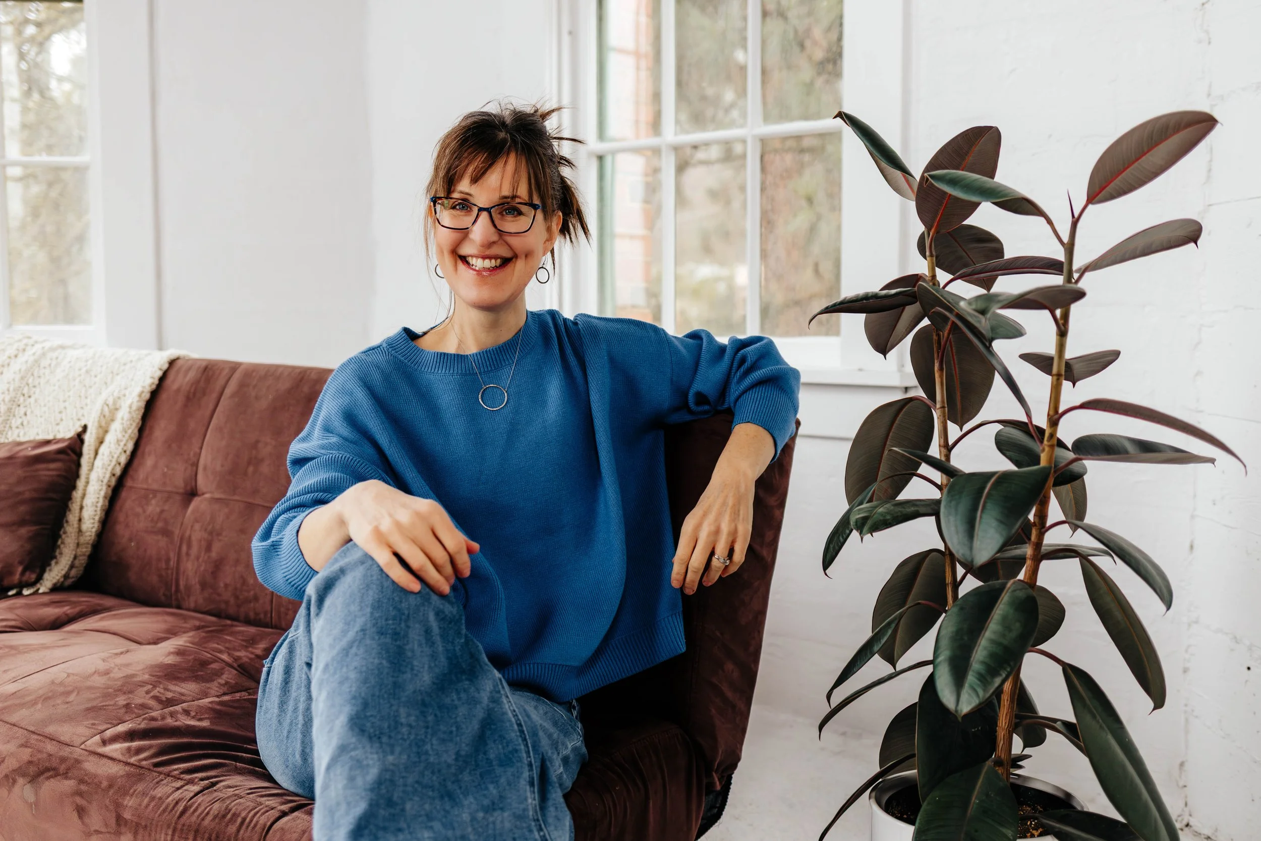 Woman with blonde hair and tattoo wearing a black AC/DC T-shirt and ripped jeans sitting on the floor using a camera with large potted plants and a yellow wall in the background.