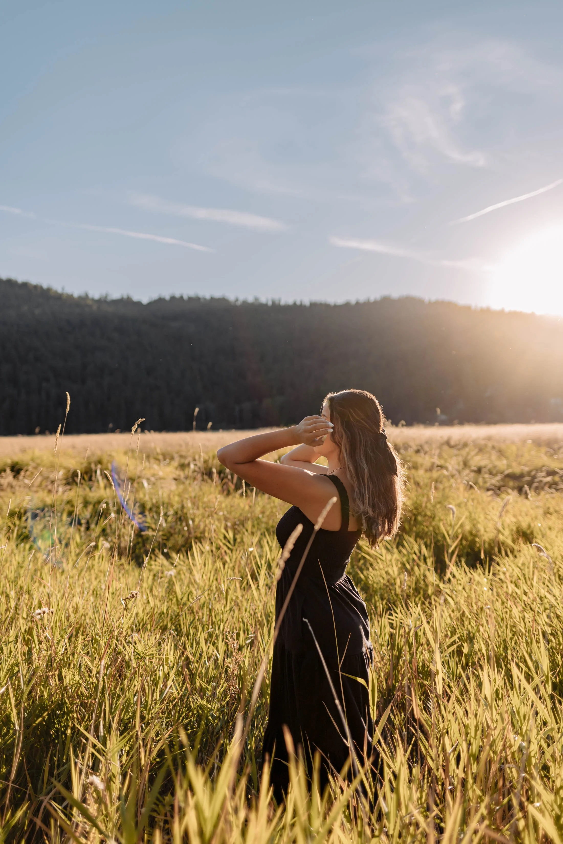 A couple embraces and smiles at each other in a green outdoor field with trees and a lake in the background during sunset.