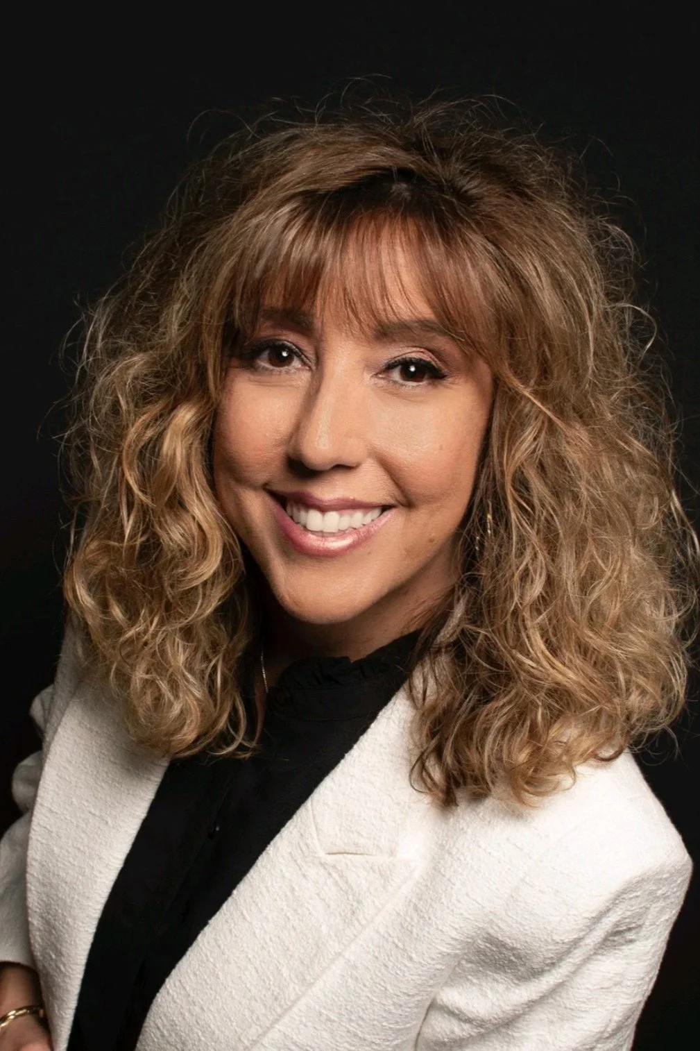 A woman with curly blonde hair, smiling, dressed in a black top and white blazer against a dark background.