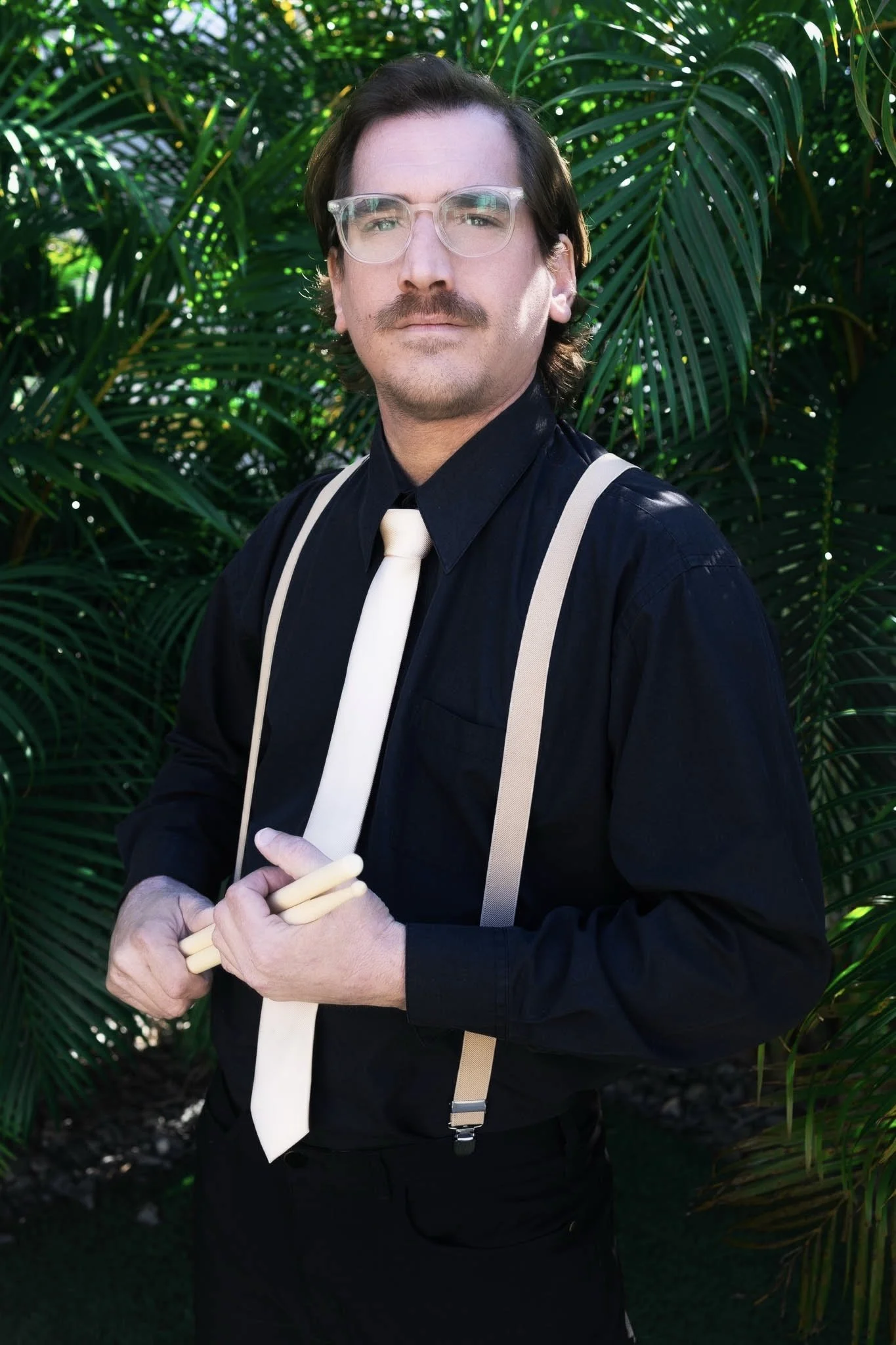 A man with shoulder-length dark hair, wearing clear glasses, a black shirt with a white tie, beige suspenders, and holding white chopsticks, standing in front of lush green tropical plants.