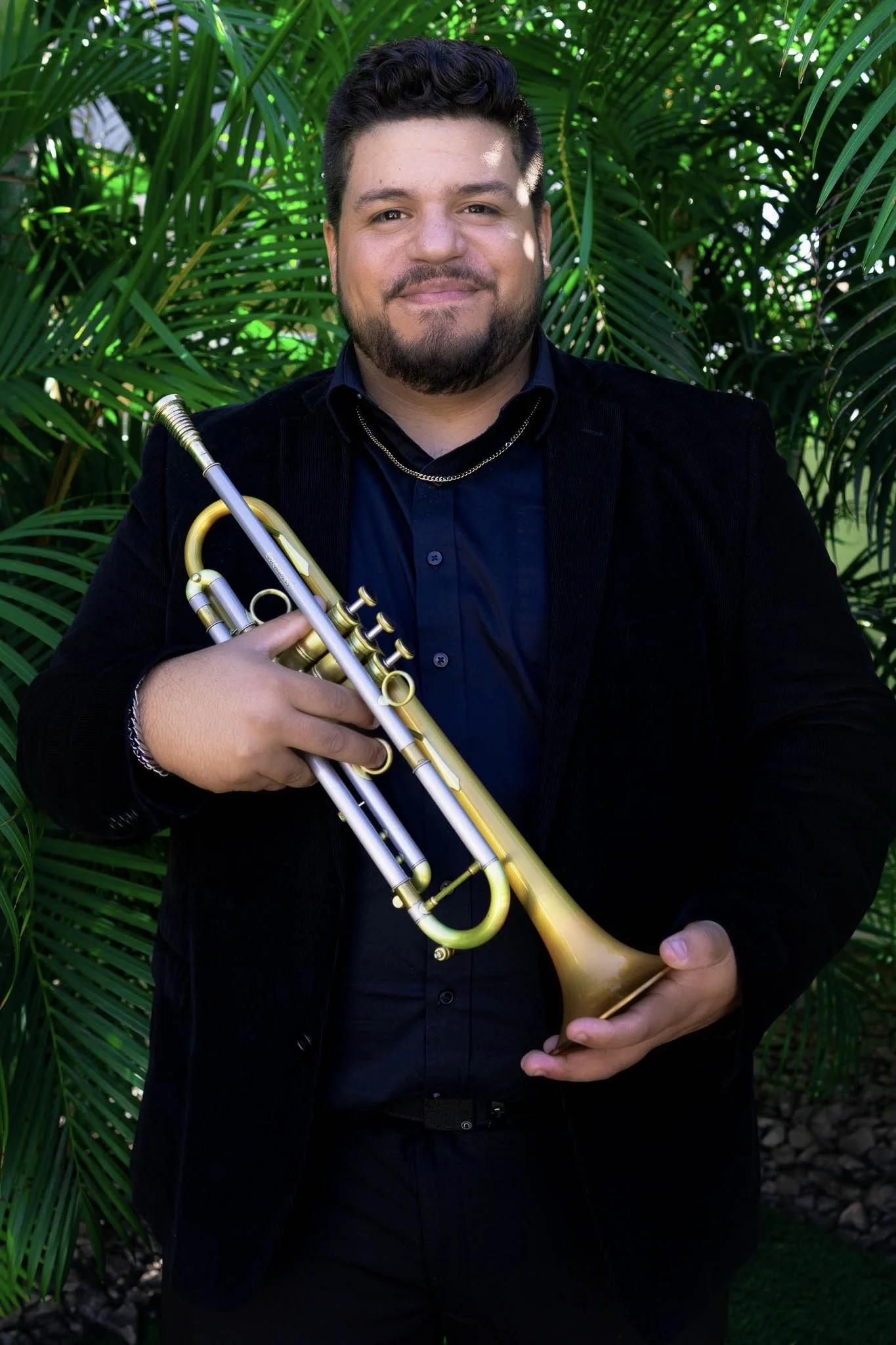 A man with dark hair and a beard playing a silver trumpet in a dimly lit room with purple and pink lighting, surrounded by musical equipment and decorative wall sconces.