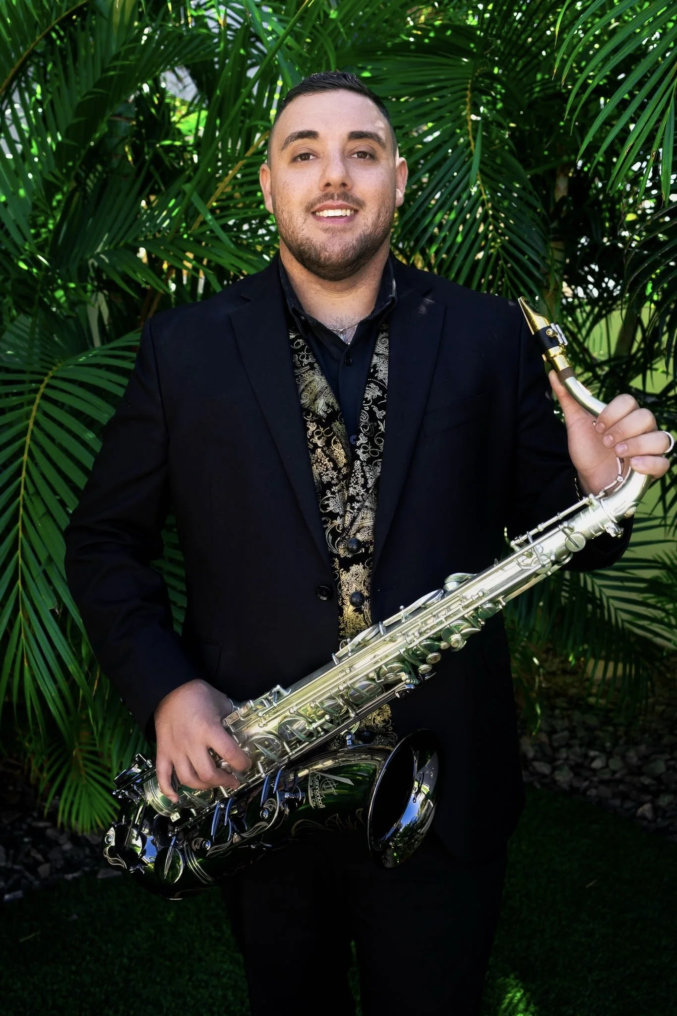 A man in a black suit holding a saxophone in front of green leafy plants.