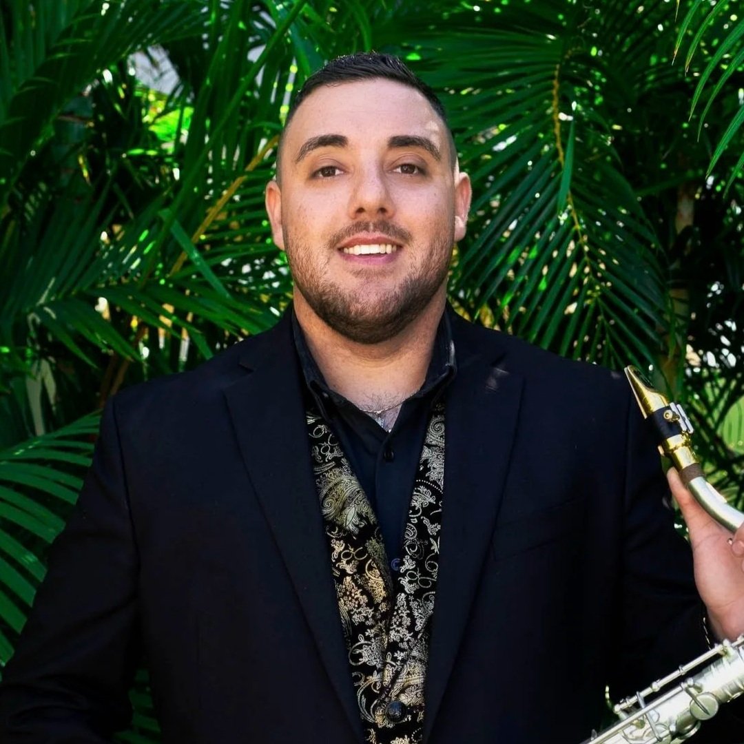 Portrait of a smiling man with short dark hair, light beard, wearing a dark shirt, with blurred shelves in the background.