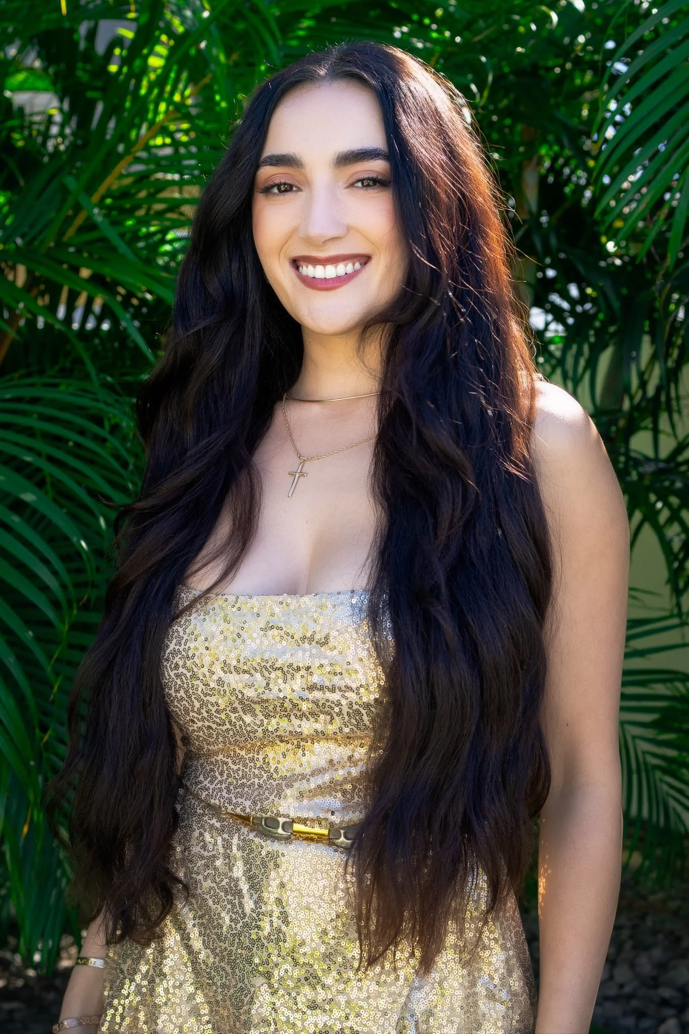 A young woman with long dark wavy hair smiling in front of lush green foliage, wearing a gold sequined dress and gold jewelry.