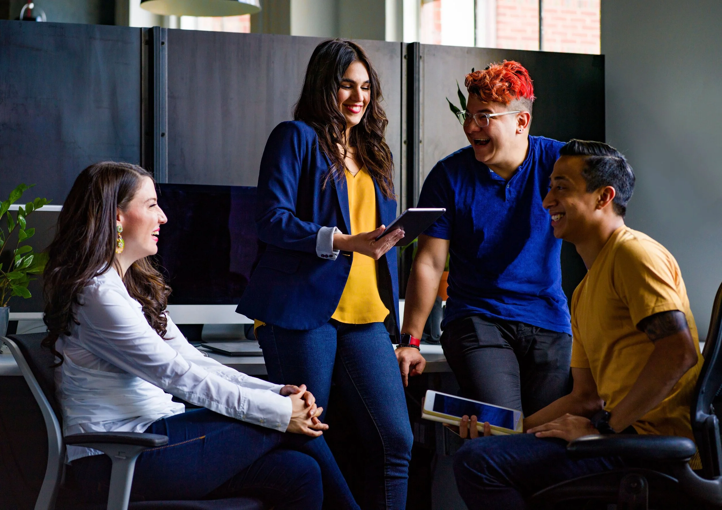 Four diverse young adults in an office, engaging in a cheerful discussion, with one woman standing and holding a tablet while the others are seated and smiling.