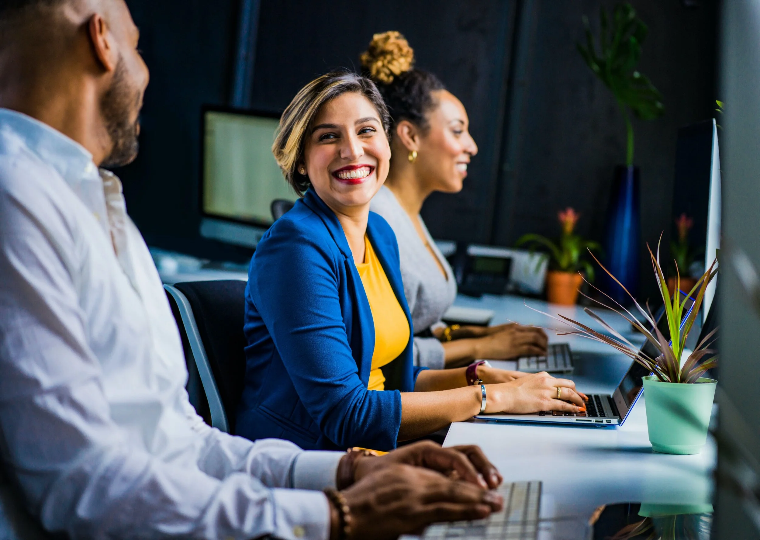 Three diverse coworkers working at computers in an office, with one woman smiling and looking at the camera.