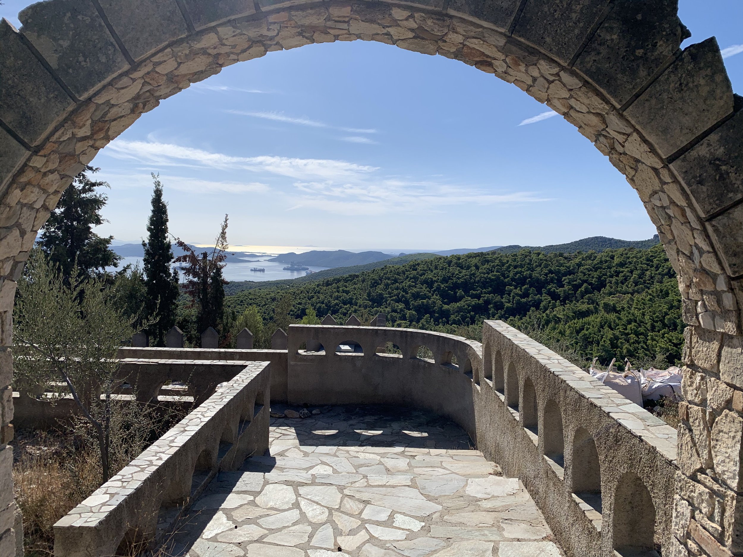 Beautiful cobble stone walking path / bridge leading under an archway to a scenic hillside view covered in green trees with a sea in the distance on a Greek Island