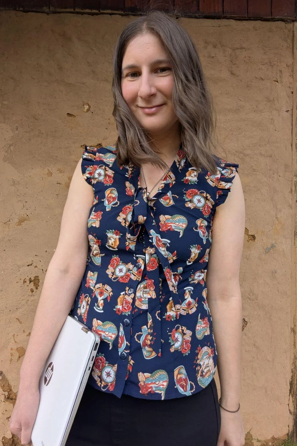 Sarah holding a laptop wearing a summer top standing in front of a brown wall