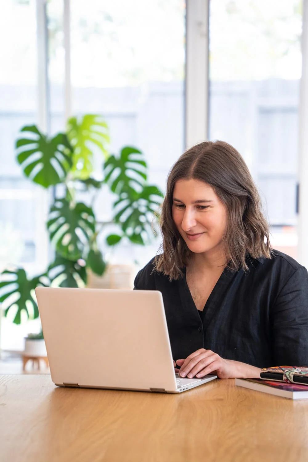 A website copywriter from Melbourne with brown hair, wearing black clothes, smiles while working on a laptop at a wooden table. A large green plant is visible in the background