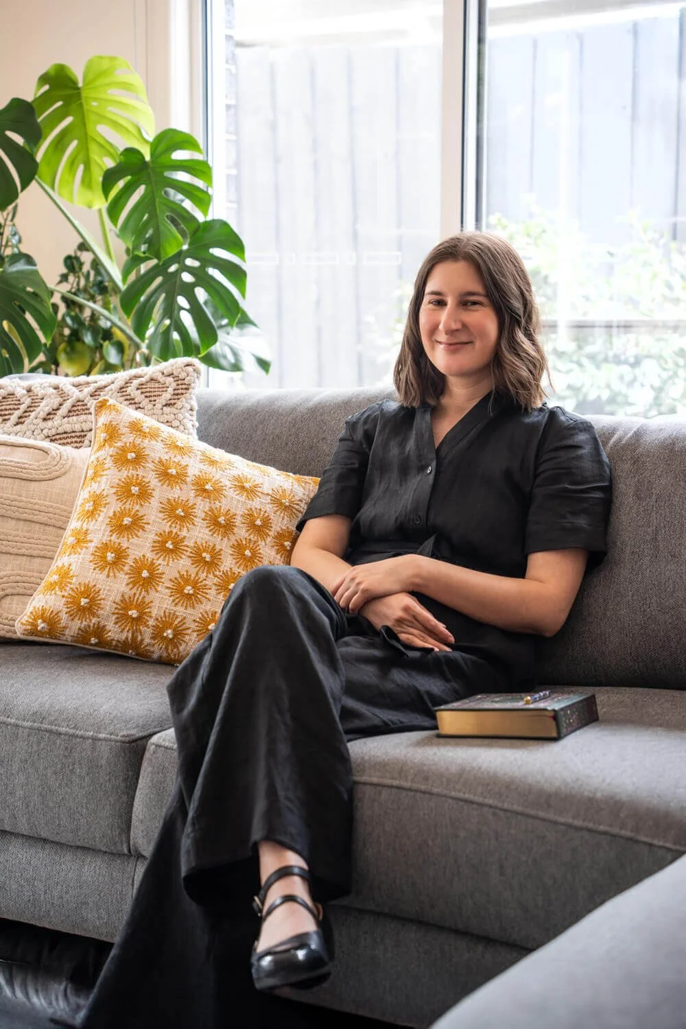 Hire copywriter Sarah Marriott from Melbourne sitting on a grey couch with a notebook and pen with green plants and yellow cushions in the background