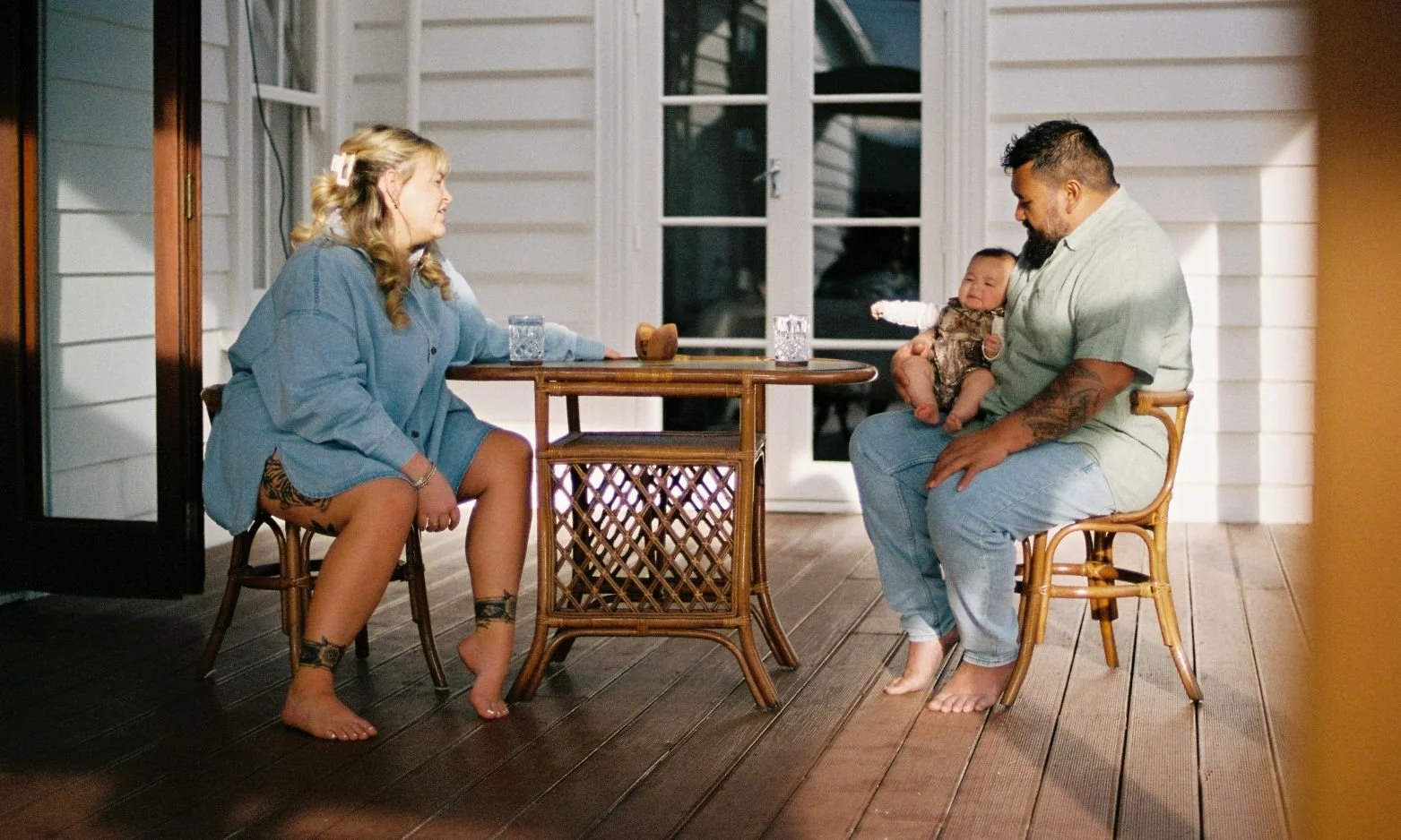 A family photo taken by Moa Raud, in a natural setting on a deck at a table, featuring a mother, father and baby